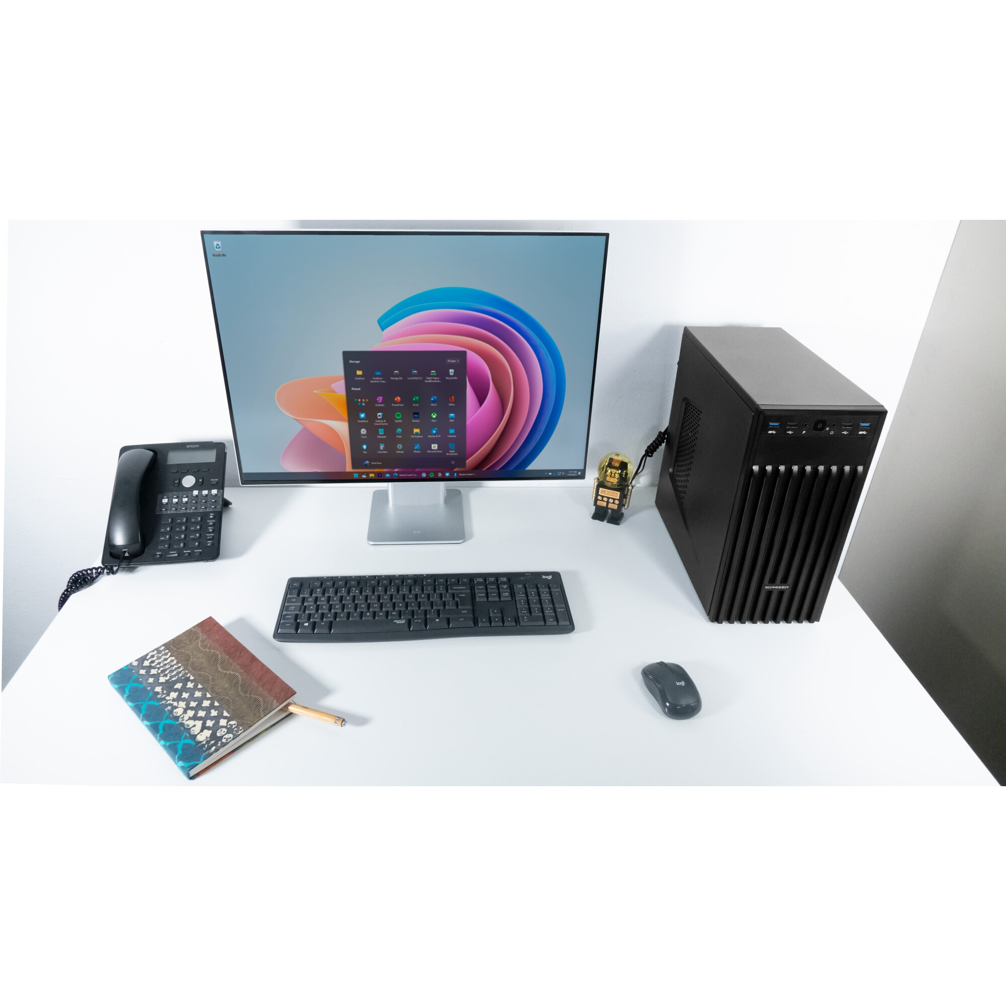 Biurko z monitorem, klawiaturą i wieżą komputera. Telefon i notatnik po lewej stronie. Białe tło.