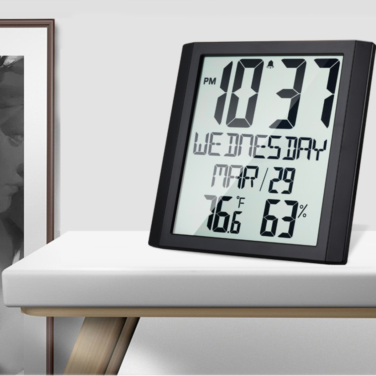 Schwarze Digitaluhr, die Zeit, Datum und Temperatur anzeigt. Auf einem weißen Tisch, mit einem Bilderrahmen.