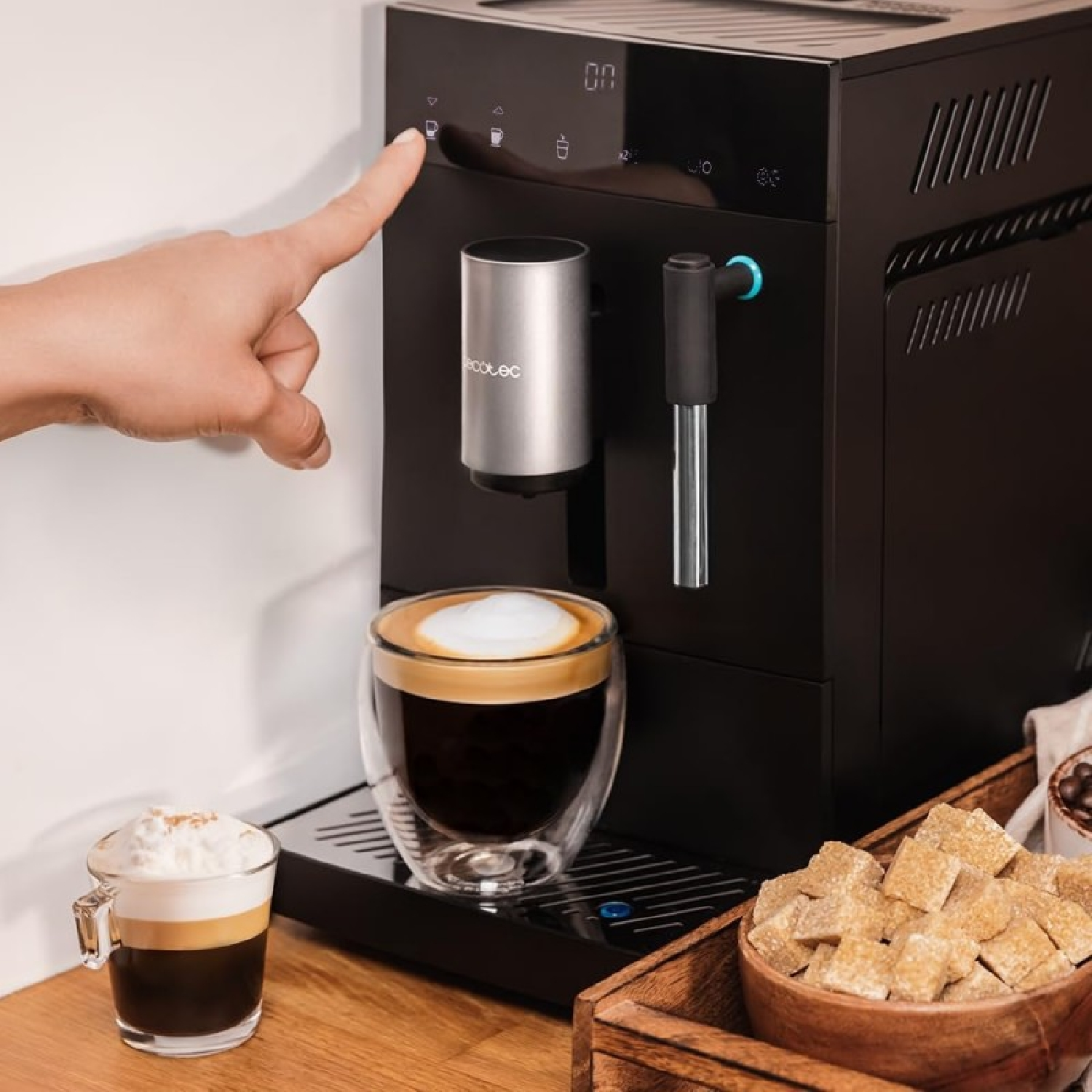 Un dito preme un pulsante della macchina del caffè. Un bicchiere di caffè e zollette di zucchero in primo piano.