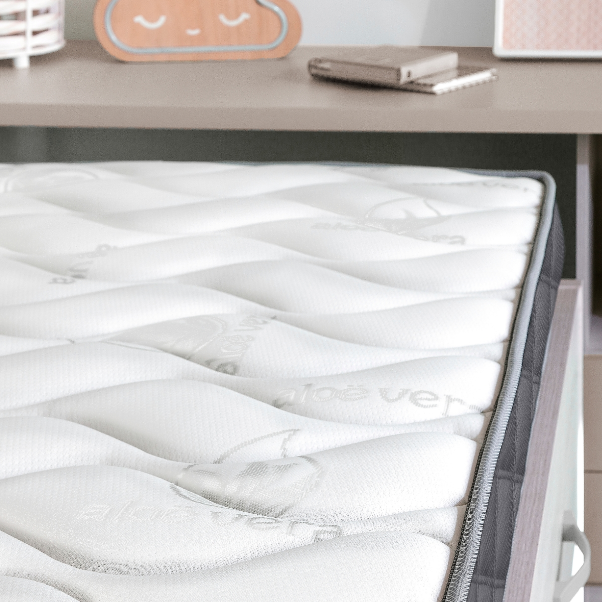 Colchón blanco con borde gris sobre un marco de cama. Objetos en una estantería al fondo.
