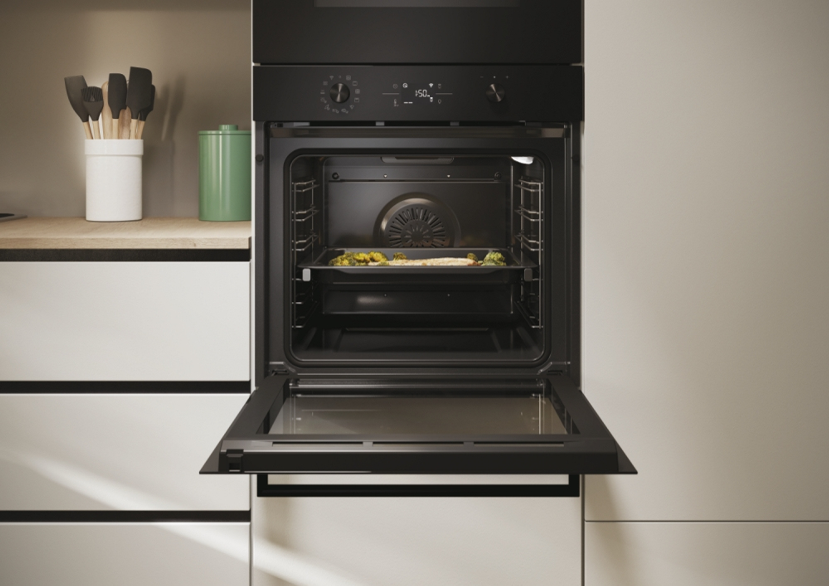 Horno negro con puerta abierta, bandeja con verduras dentro. Encimera de madera, utensilios visibles.