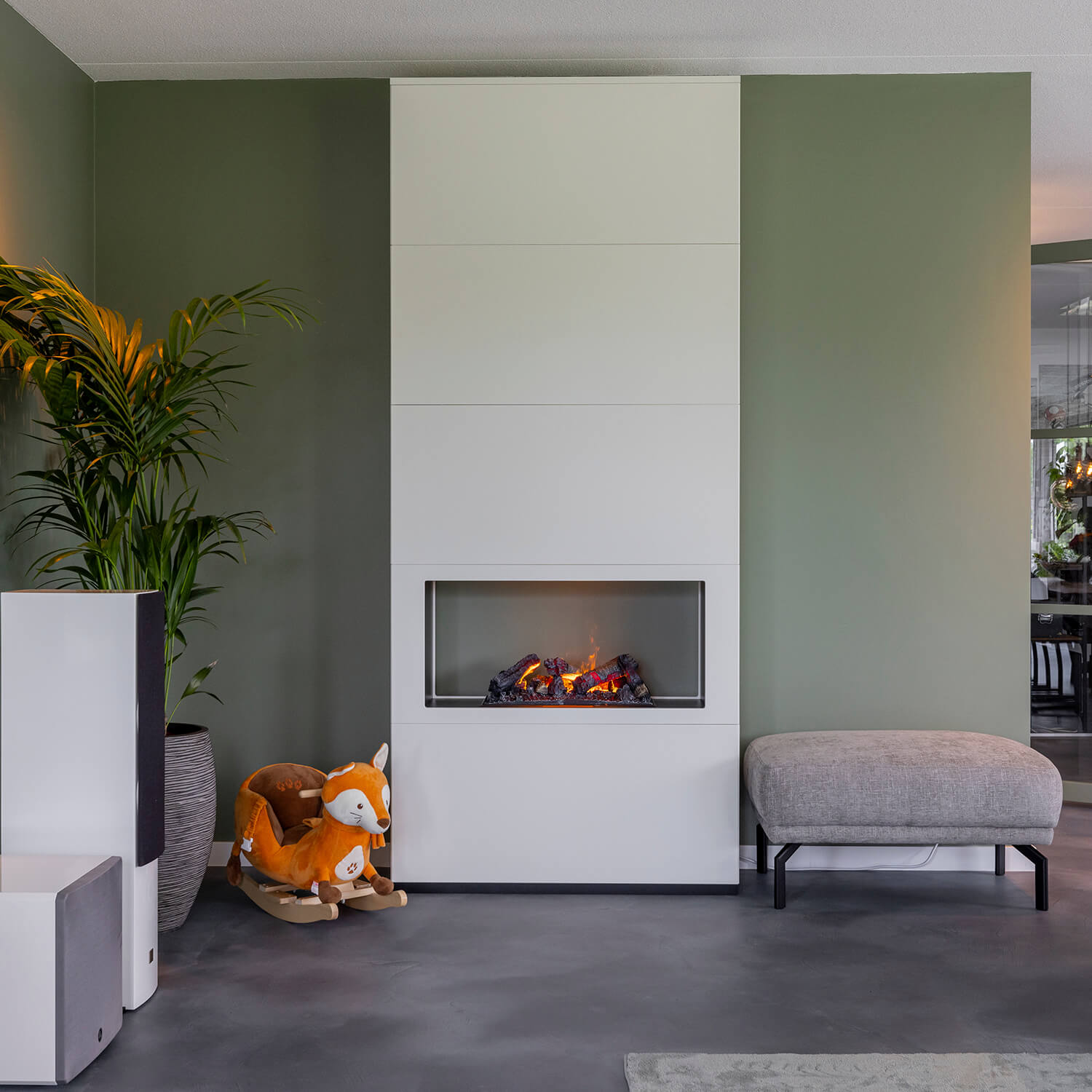 Een moderne woonkamer met een witte open haard. Een schommelvos, een plant in pot en een grijze bank staan voor de groene muur.