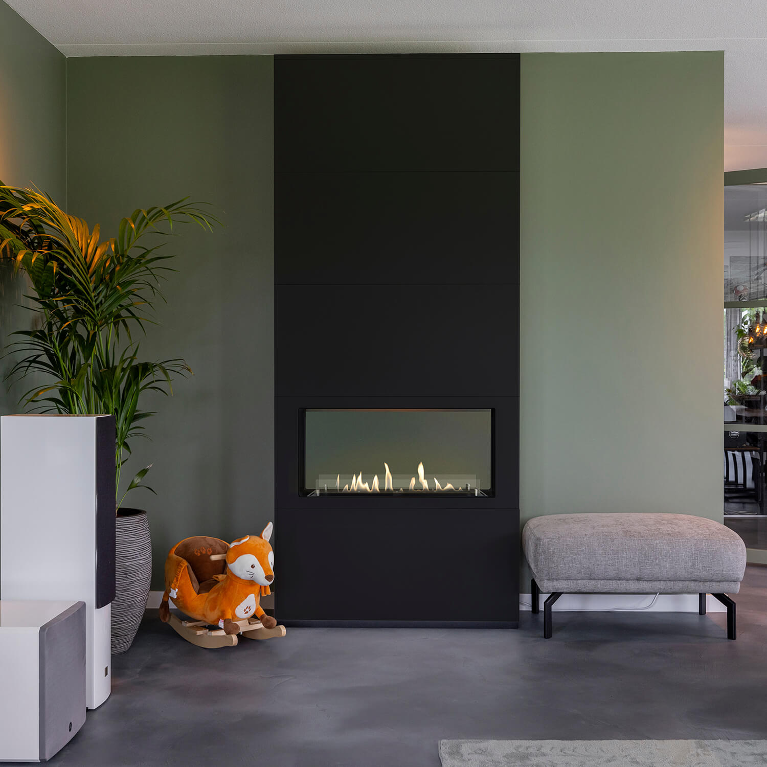 Woonkamer met een zwarte open haard, een vossenwip en een grijze bank. Groene muren en een potplant zorgen voor kleur.
