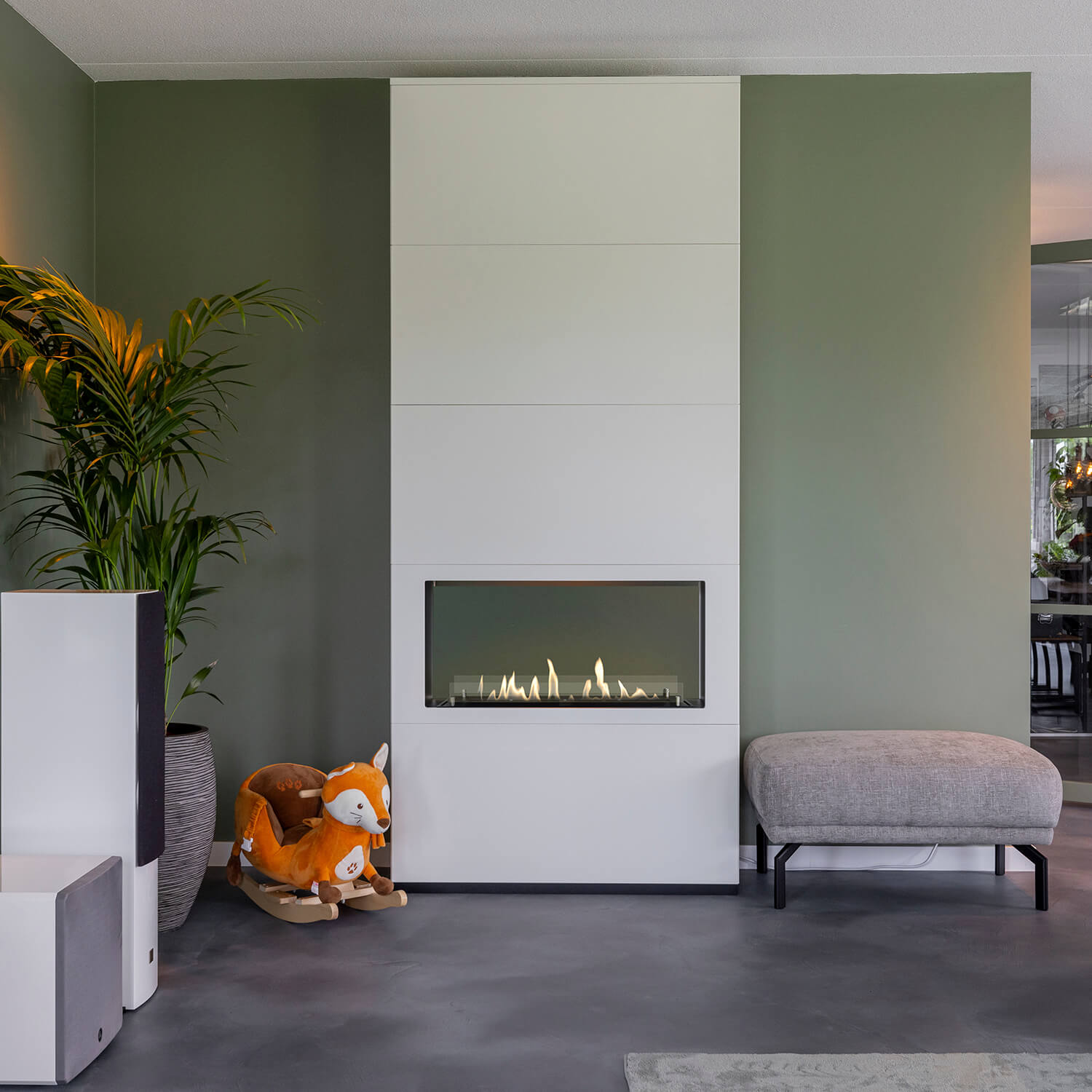 Een moderne woonkamer met een witte open haard, een oranje vos schommel, een plant en een grijze bank. Groene muur.