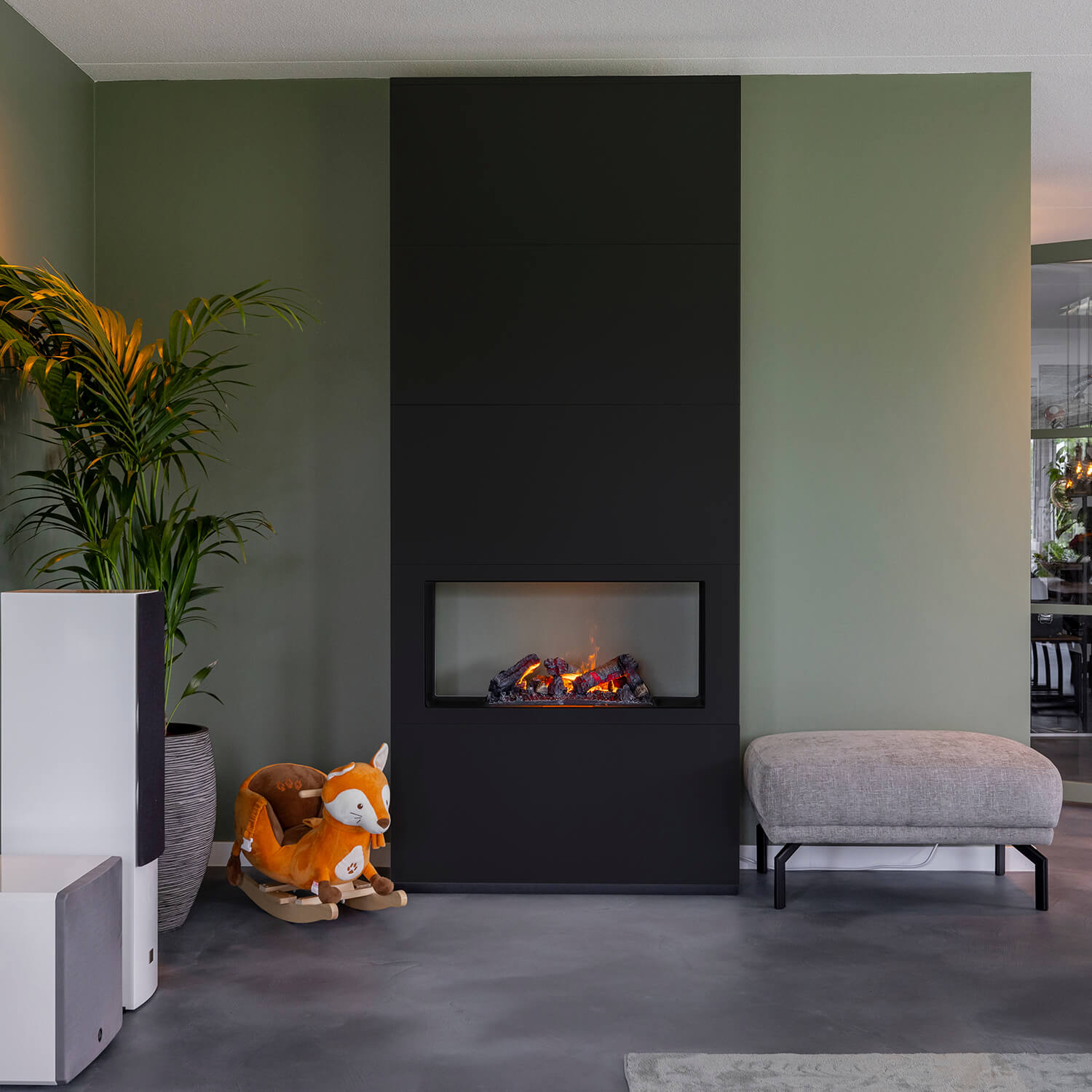 Woonkamer met open haard, plant, schommelvos en grijze poef. Muren zijn groen en zwart.