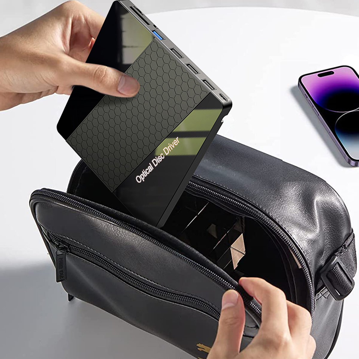 Eine Person legt einen schwarzen optischen Laufwerk in eine schwarze Tasche. Ein Telefon ist in der Nähe.