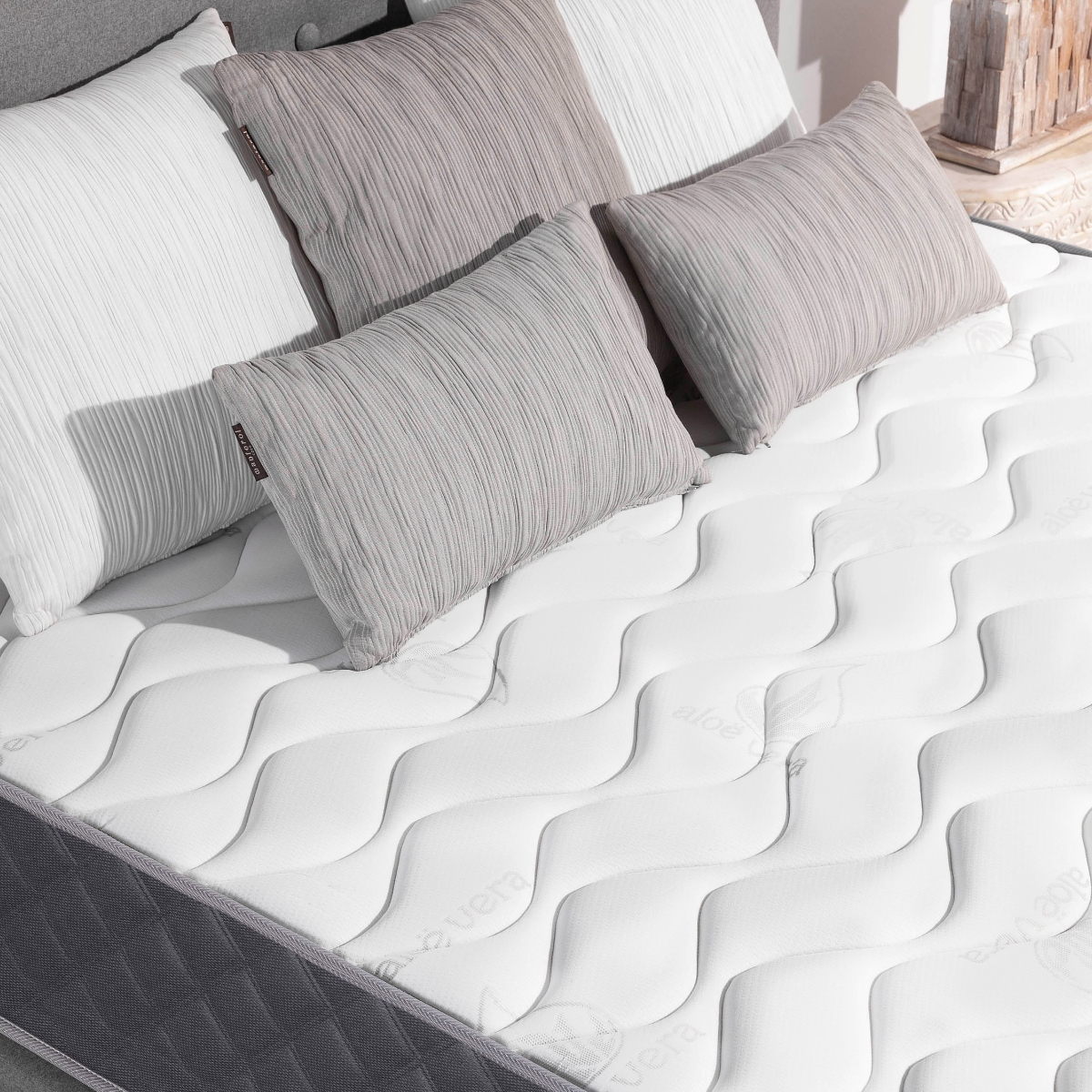 Colchón blanco con borde gris y almohadas. El colchón tiene una superficie ondulada y varias almohadas encima.