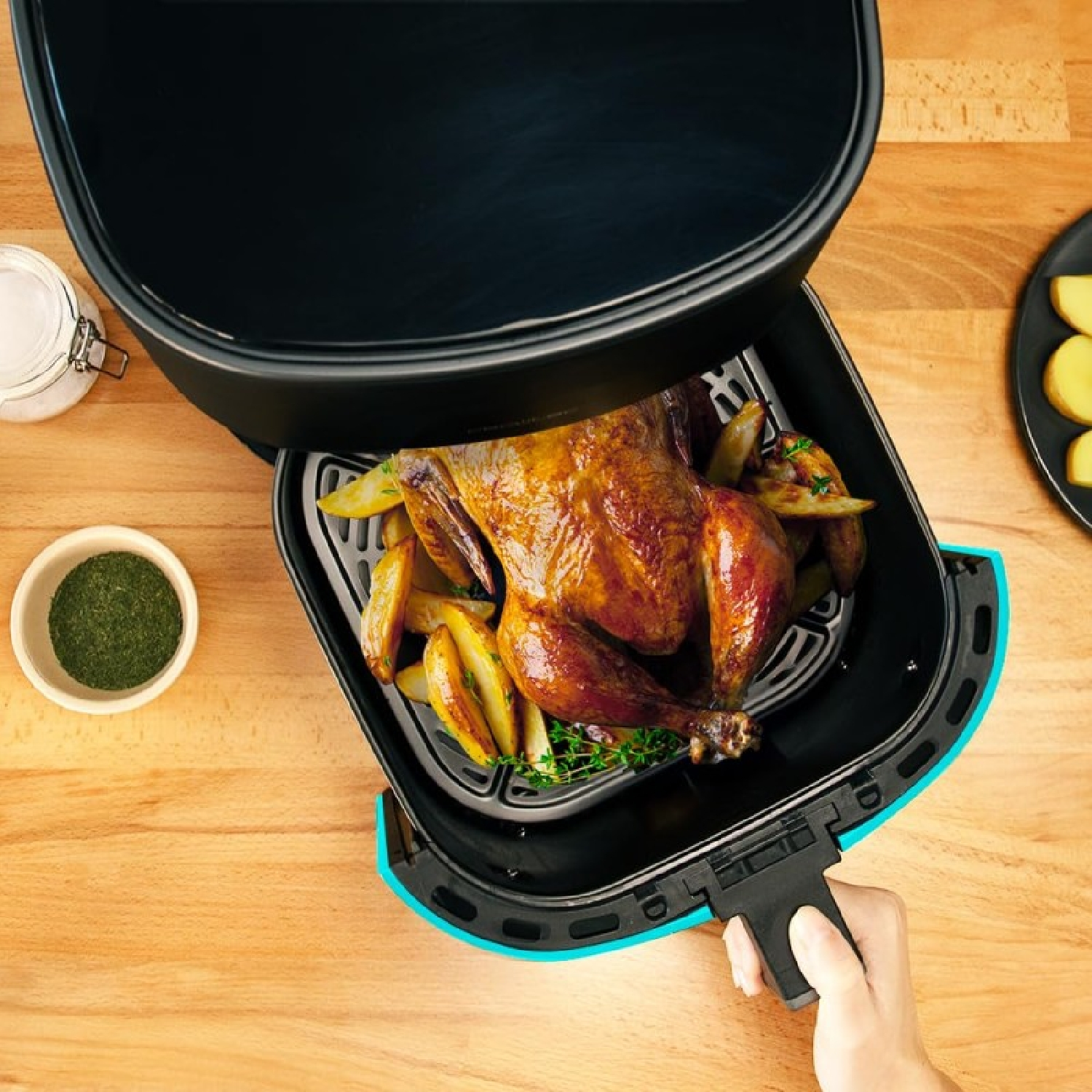 Freidora de aire con pollo cocinado y gajos de patata dentro, una mano sujetando el mango. Sobre una mesa.