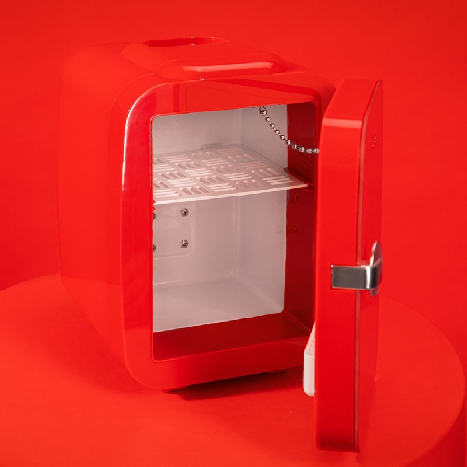 Un mini-frigorífico rojo con la puerta abierta, revelando un interior blanco y una estantería. Sobre una mesa roja.