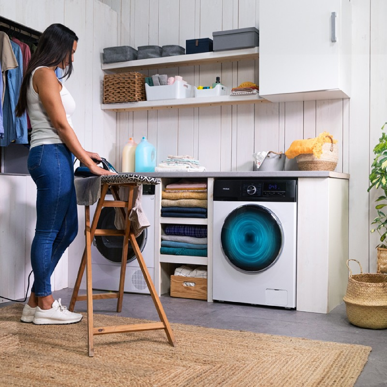 Kobieta prasująca ubrania w pralni. Jest pralka i suszarka oraz przedmioty do prania.