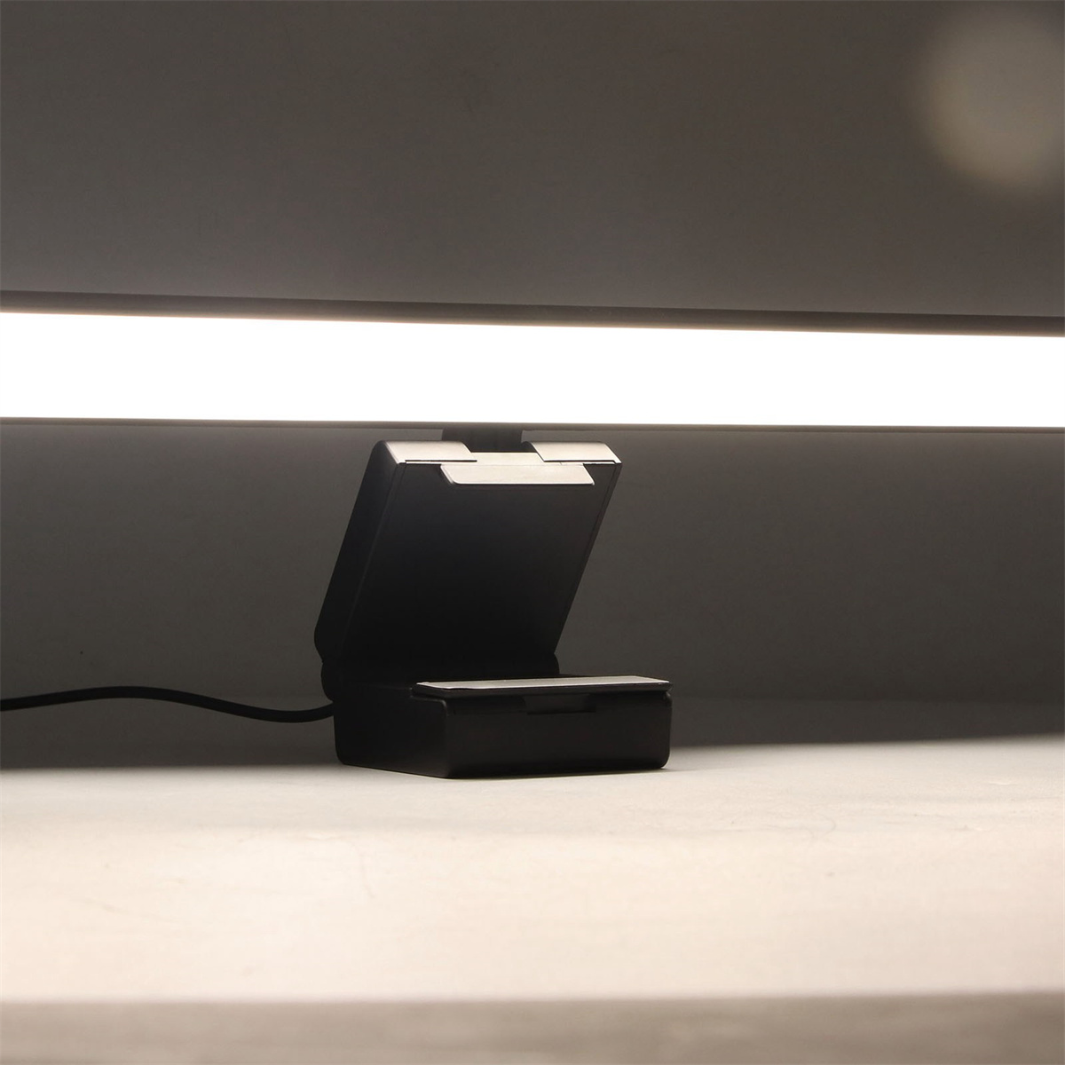 Schwarzes Gerät auf weißer Oberfläche, mit Lichtleiste darüber, in grauer Umgebung.