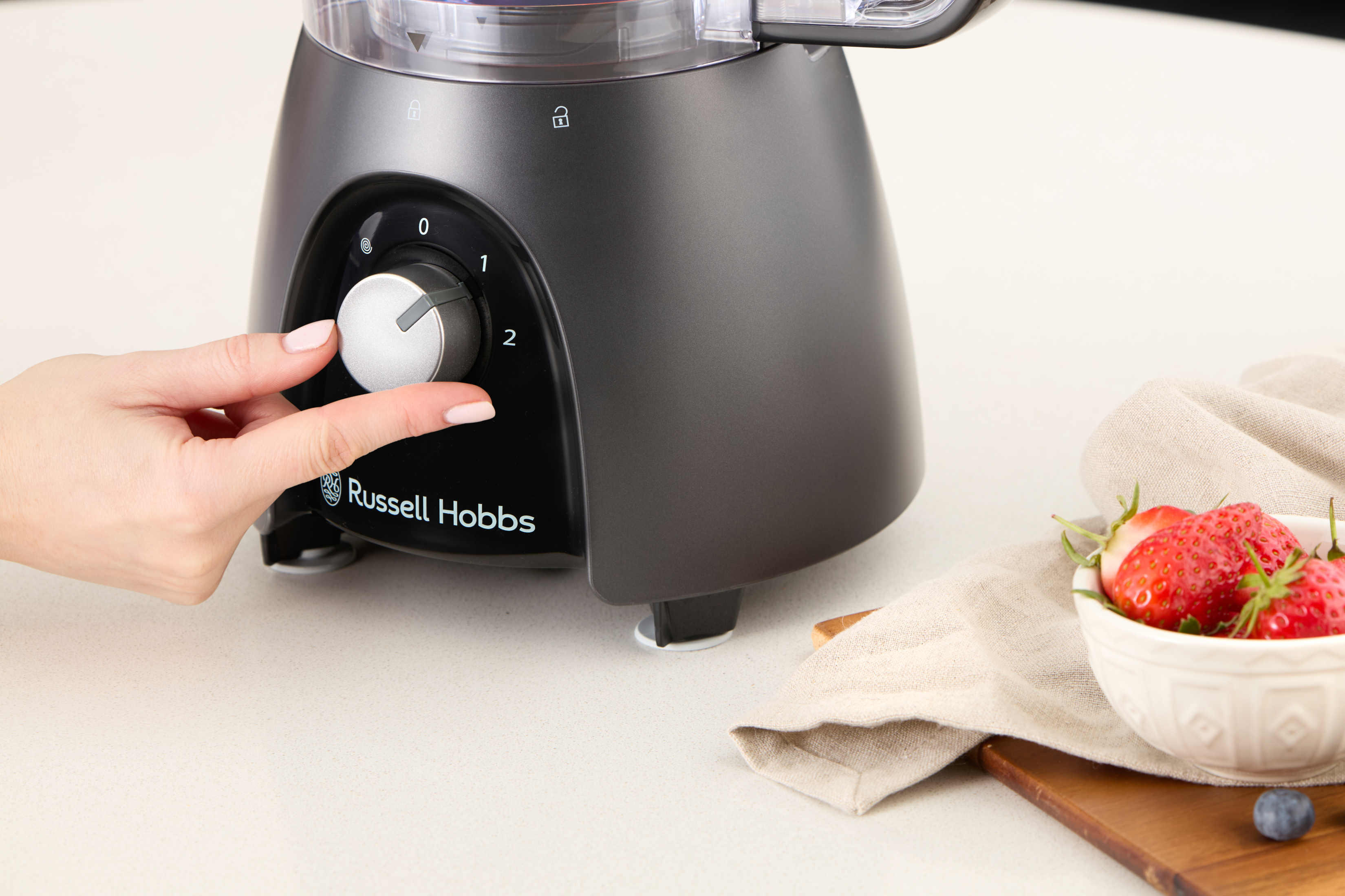 Die Hand einer Person dreht den Drehregler eines schwarzen Russell Hobbs-Zerkleinerers, Erdbeeren daneben.