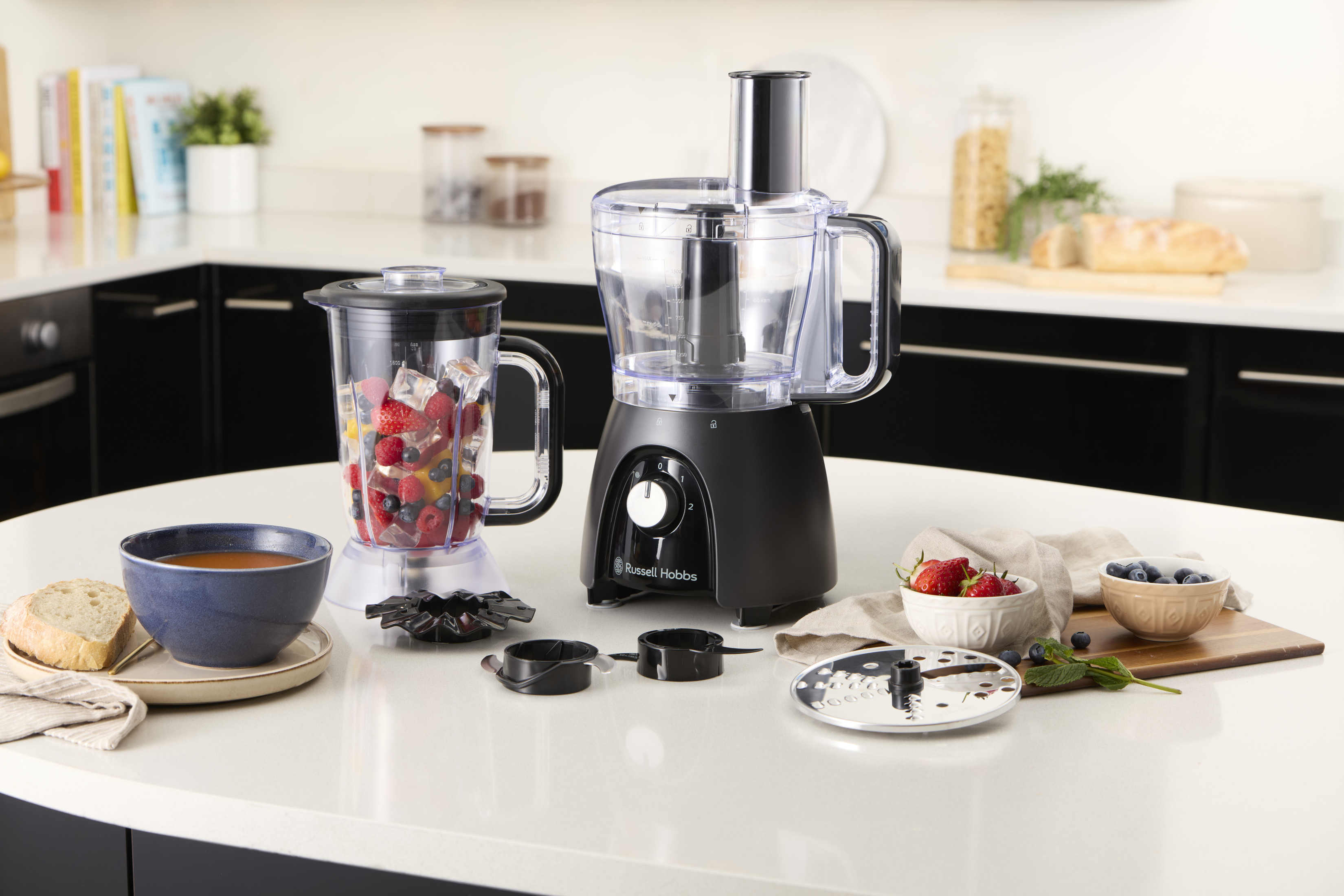Eine Küchenmaschine mit Zutaten für eine Mahlzeit. Enthält einen Mixer, eine Schüssel mit Obst und eine Schüssel mit Suppe.