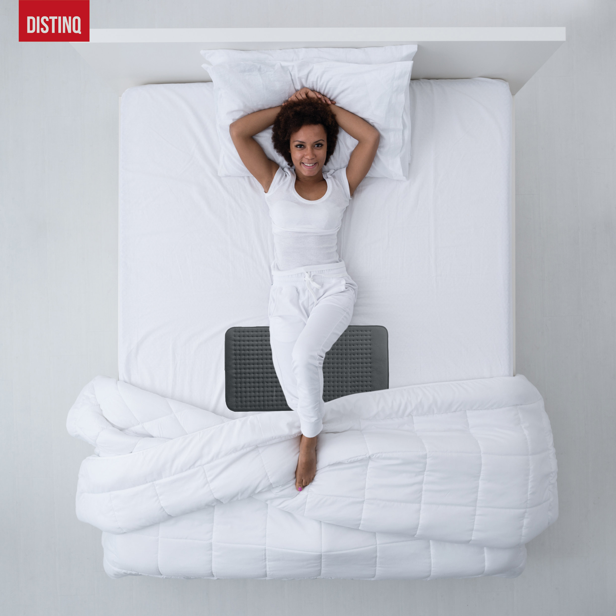 Woman lies on a bed, smiling. She wears white clothes, arms behind her head. A gray pad lies under her.