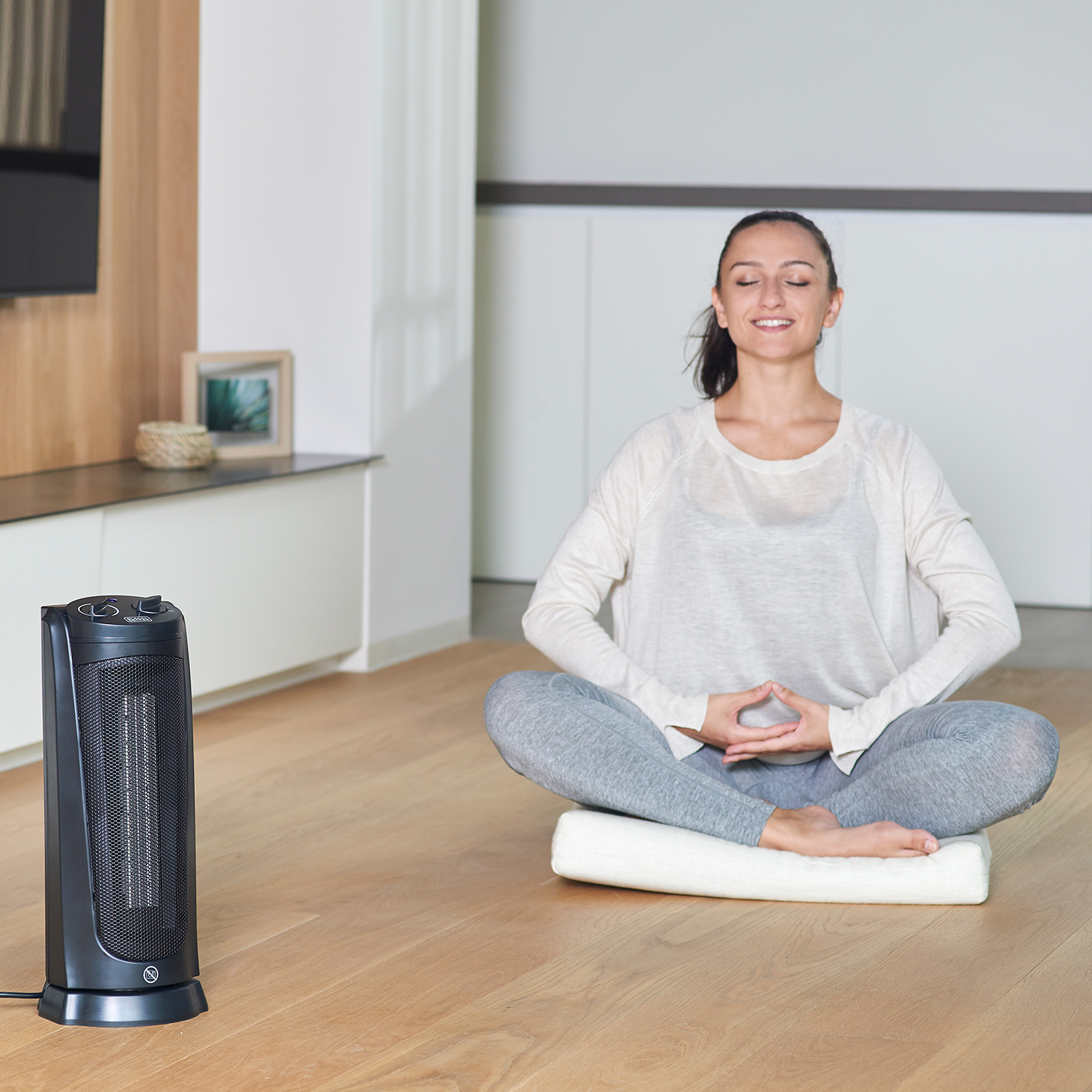 Kobieta medytuje na podłodze obok czarnego grzejnika. Ma na sobie białą koszulę i szare spodnie.
