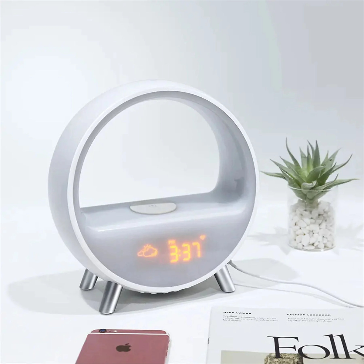 Ein weißer Wecker mit digitaler Anzeige, die 3:37 zeigt, mit einer Pflanze und einem Telefon auf dem Tisch.
