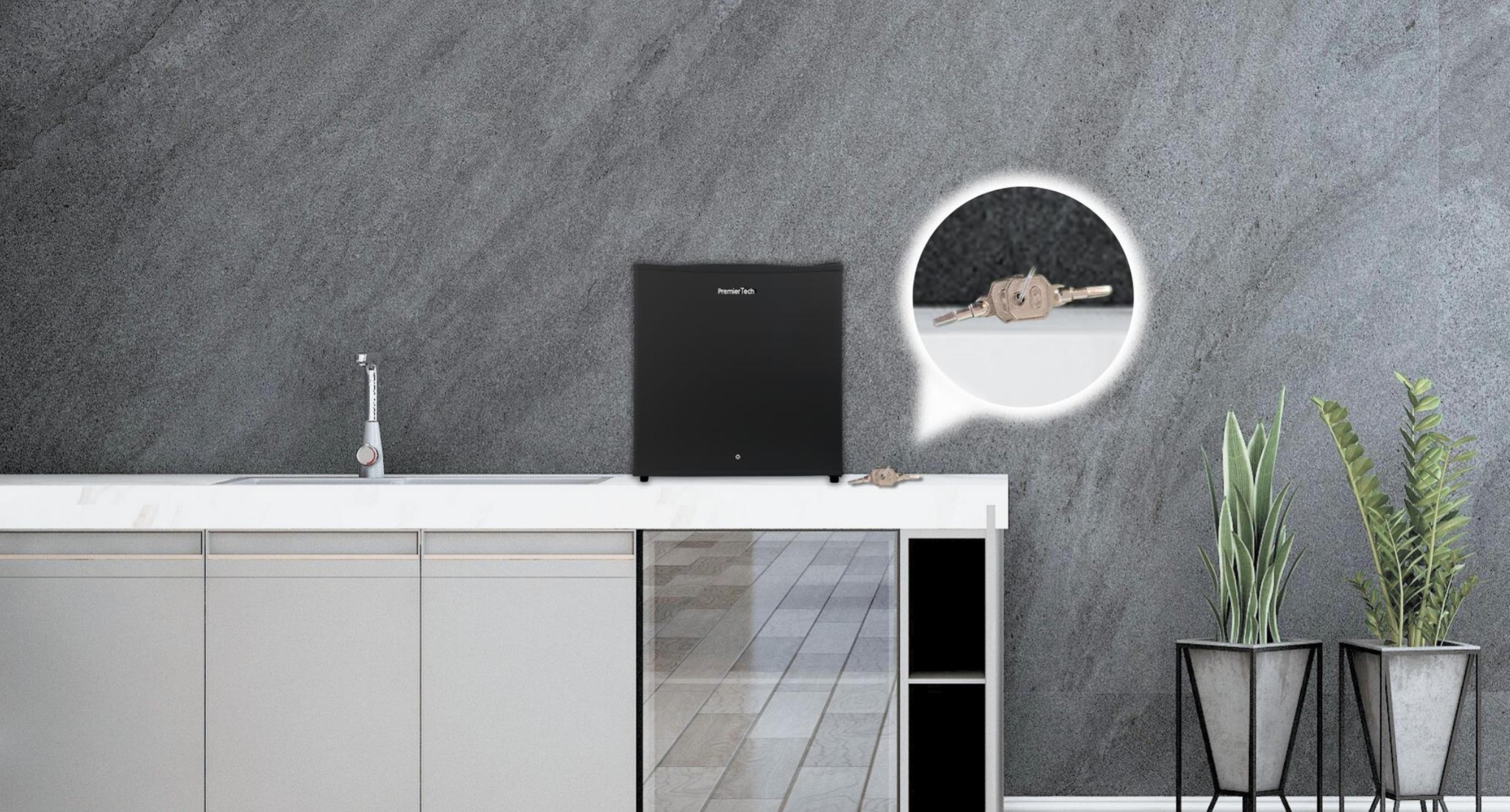 Mini-frigo PremierTech nero su un piano di lavoro bianco, con chiavi accanto e parete in pietra grigia.