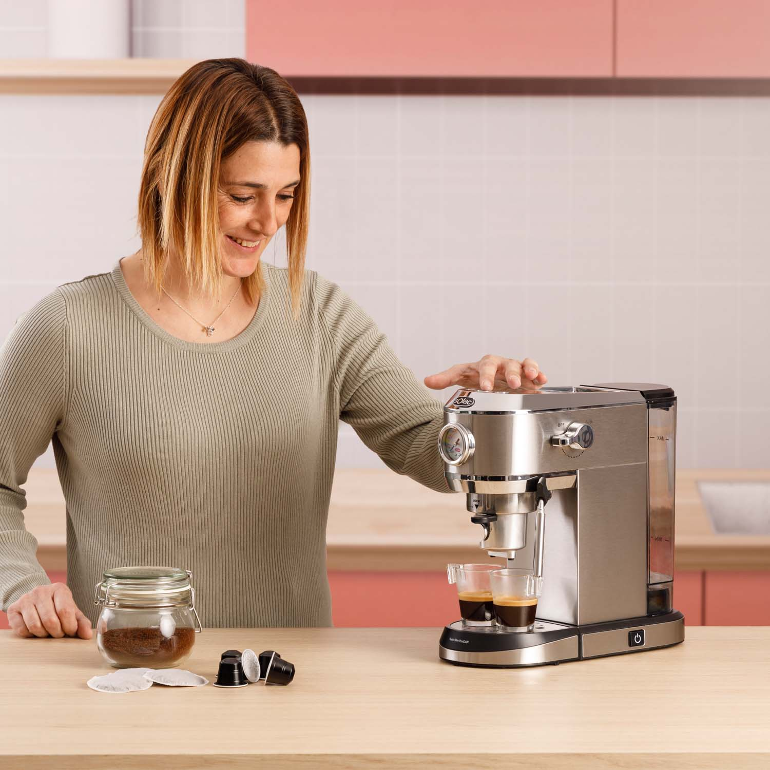 Kobieta używająca srebrnego ekspresu do kawy, z mieloną kawą i kapsułkami w pobliżu.
