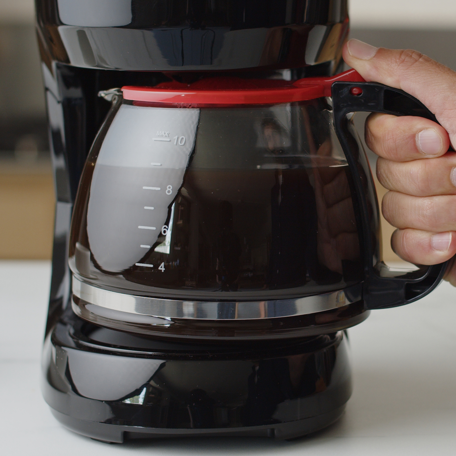 Ręka trzyma szklany dzbanek z ciemną kawą, podłączony do czarnego ekspresu do kawy.