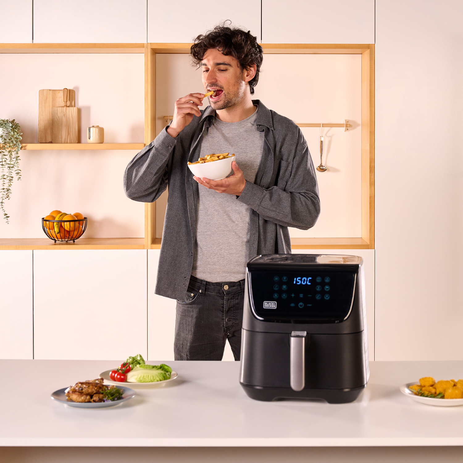 Mężczyzna je frytki stojąc obok frytkownicy. Na stole są dodatki.