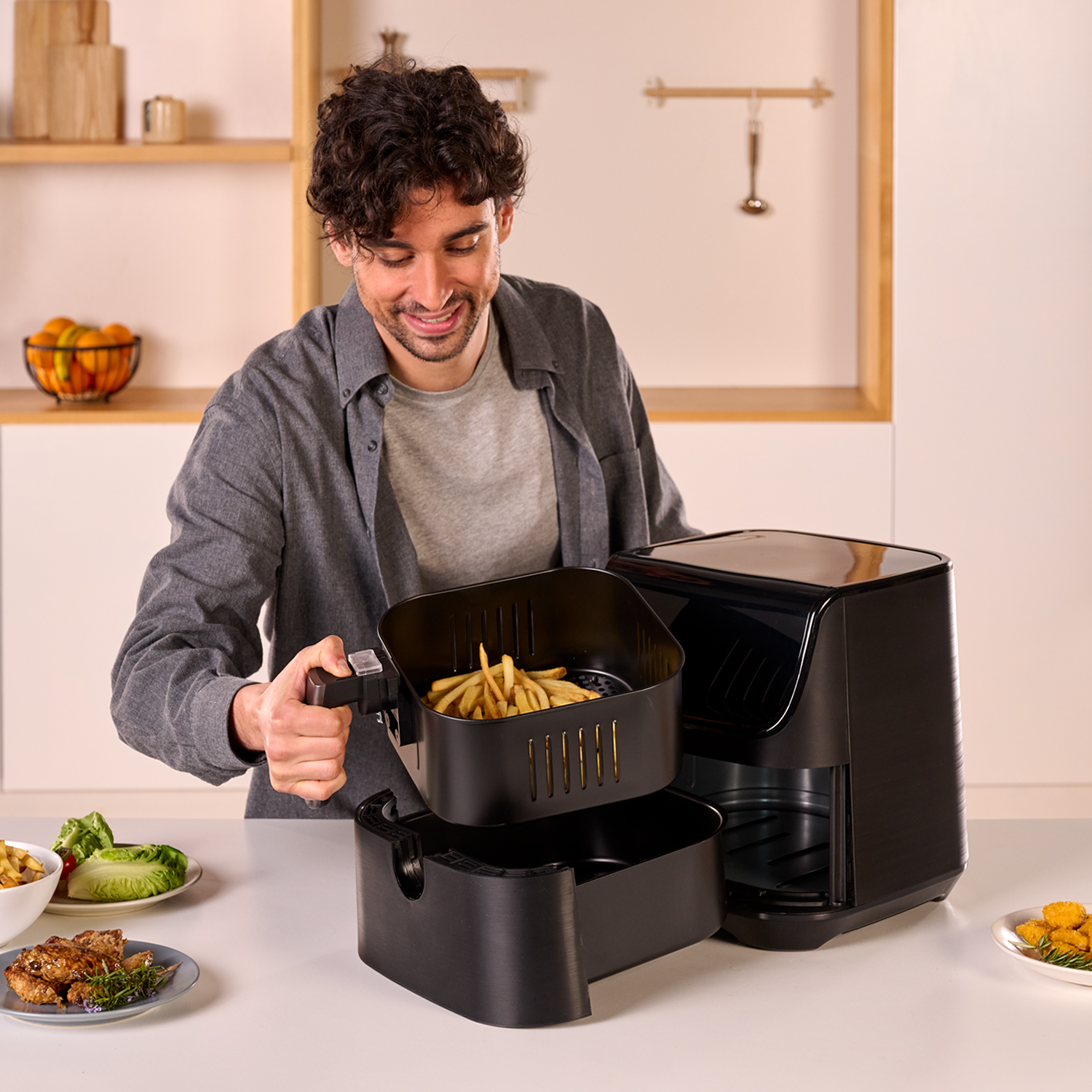 Mężczyzna wyjmuje kosz z frytkami z frytkownicy powietrznej. Na stole inne dania.