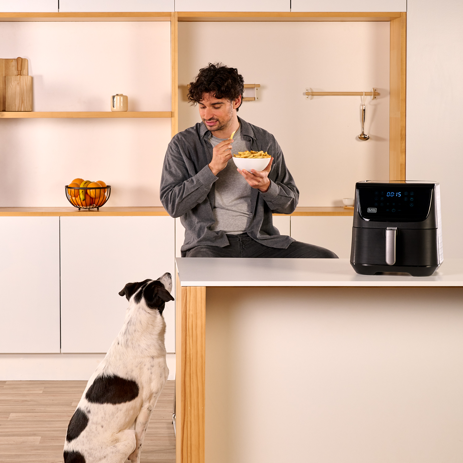 Mężczyzna je frytki z miski, pies patrzy w górę. Frytownica beztłuszczowa stoi na blacie.