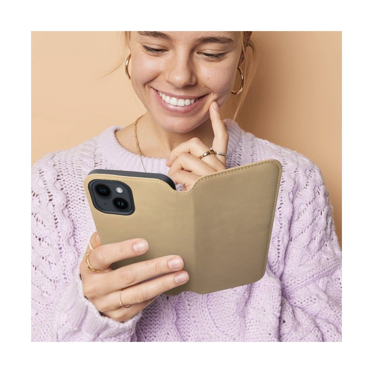 Una donna che tiene un telefono in una custodia beige, sorridente, con gioielli d'oro.