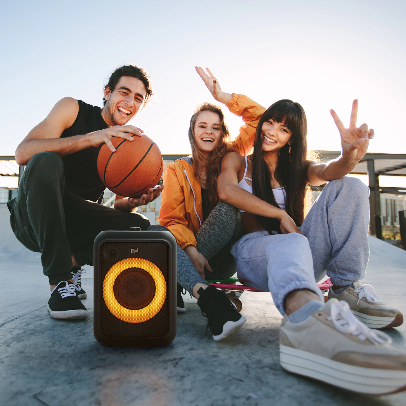 Three people with a speaker. One holds a basketball, others are happy, in a skate park.