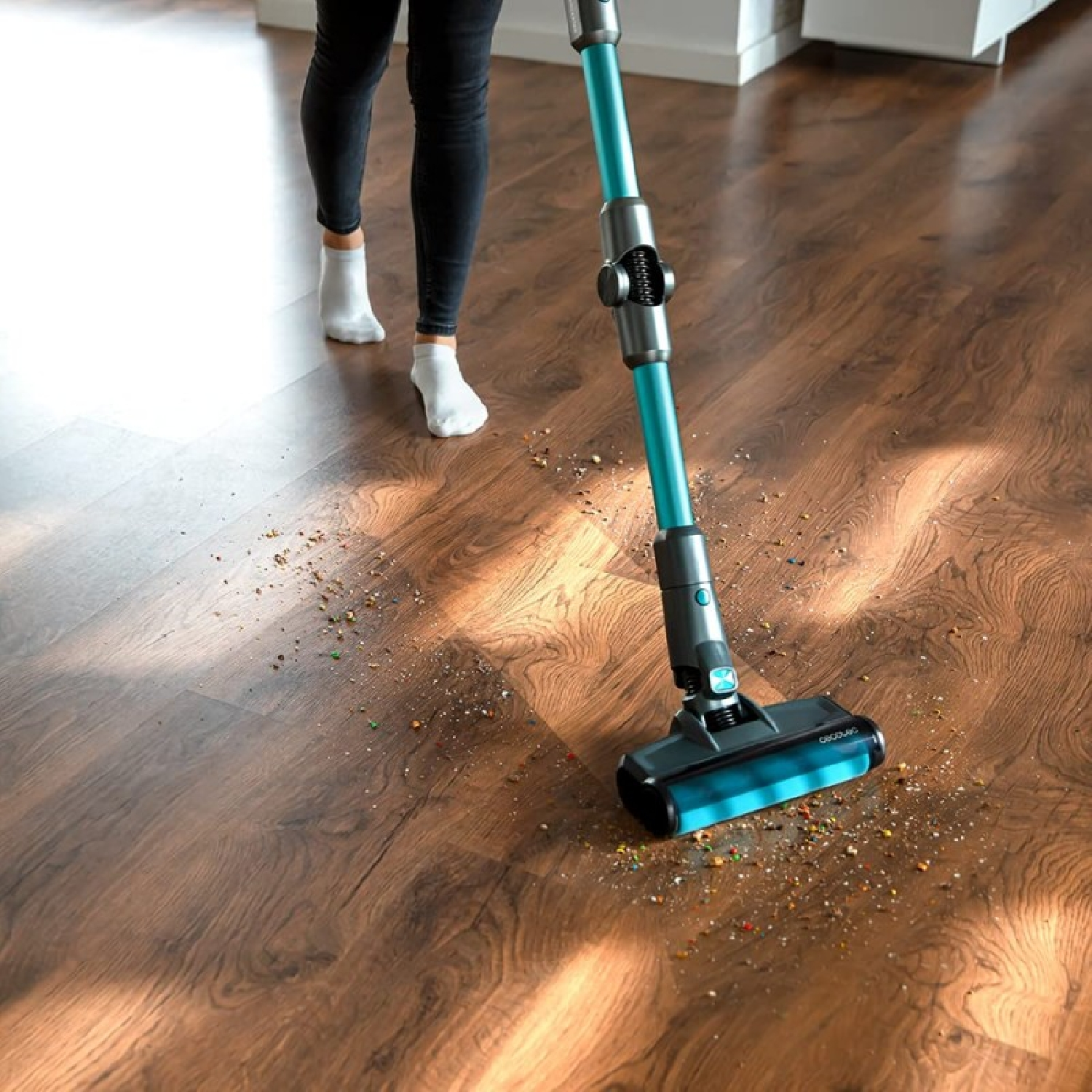 Una persona che aspira un pavimento in legno con un aspirapolvere a bastone verde acqua e grigio, spargendo detriti.
