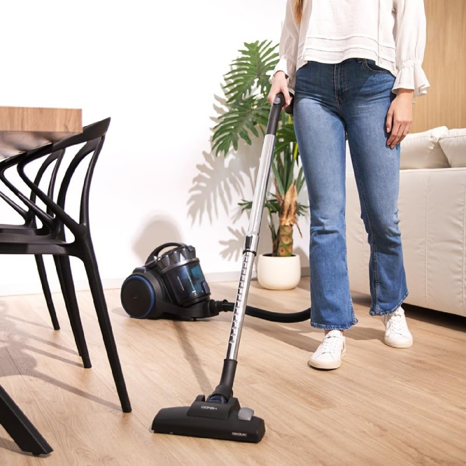 Kobieta odkurza drewnianą podłogę. Widoczny odkurzacz i czarny fotel.