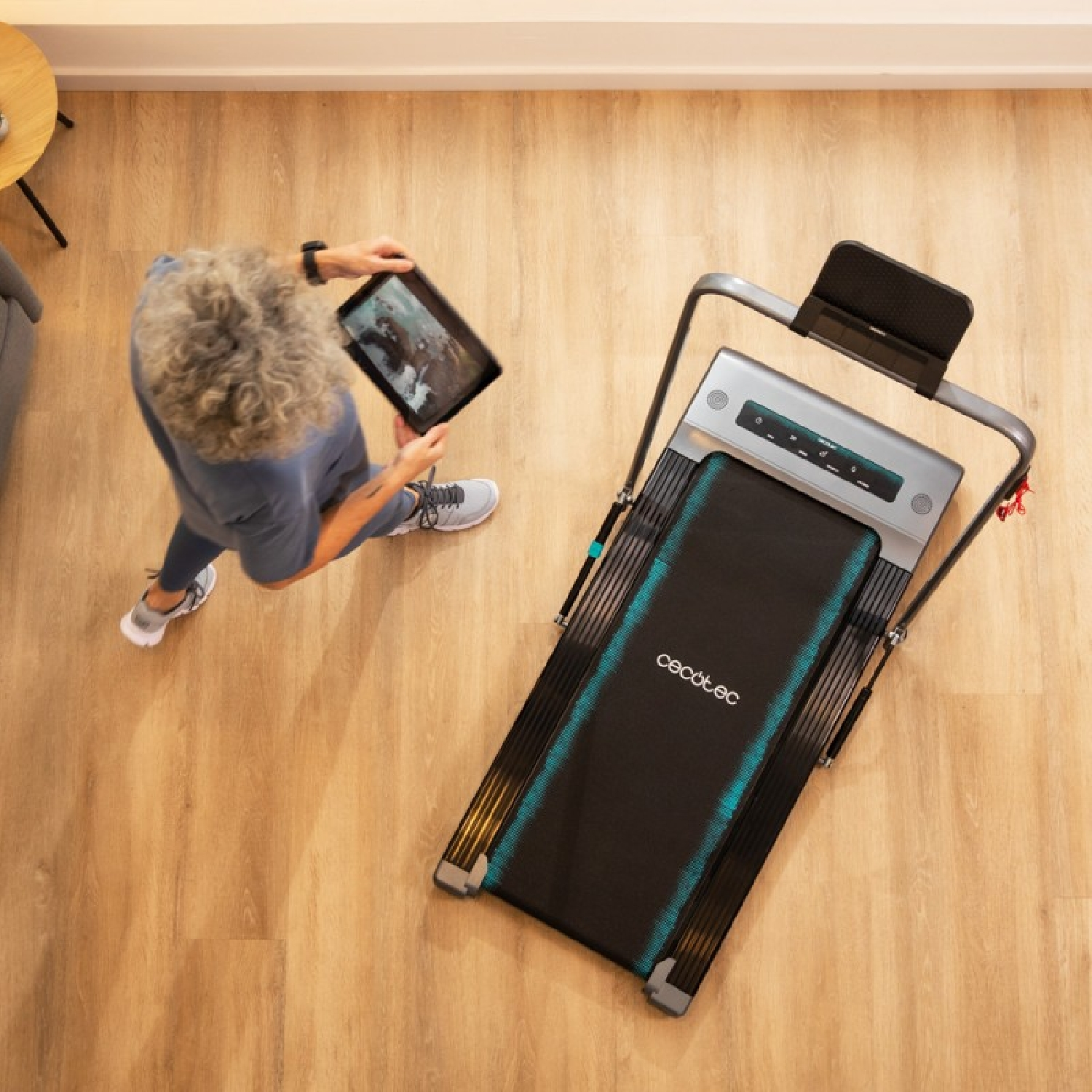 Kobieta używa tabletu przy bieżni. Bieżnia stoi na drewnianej podłodze.