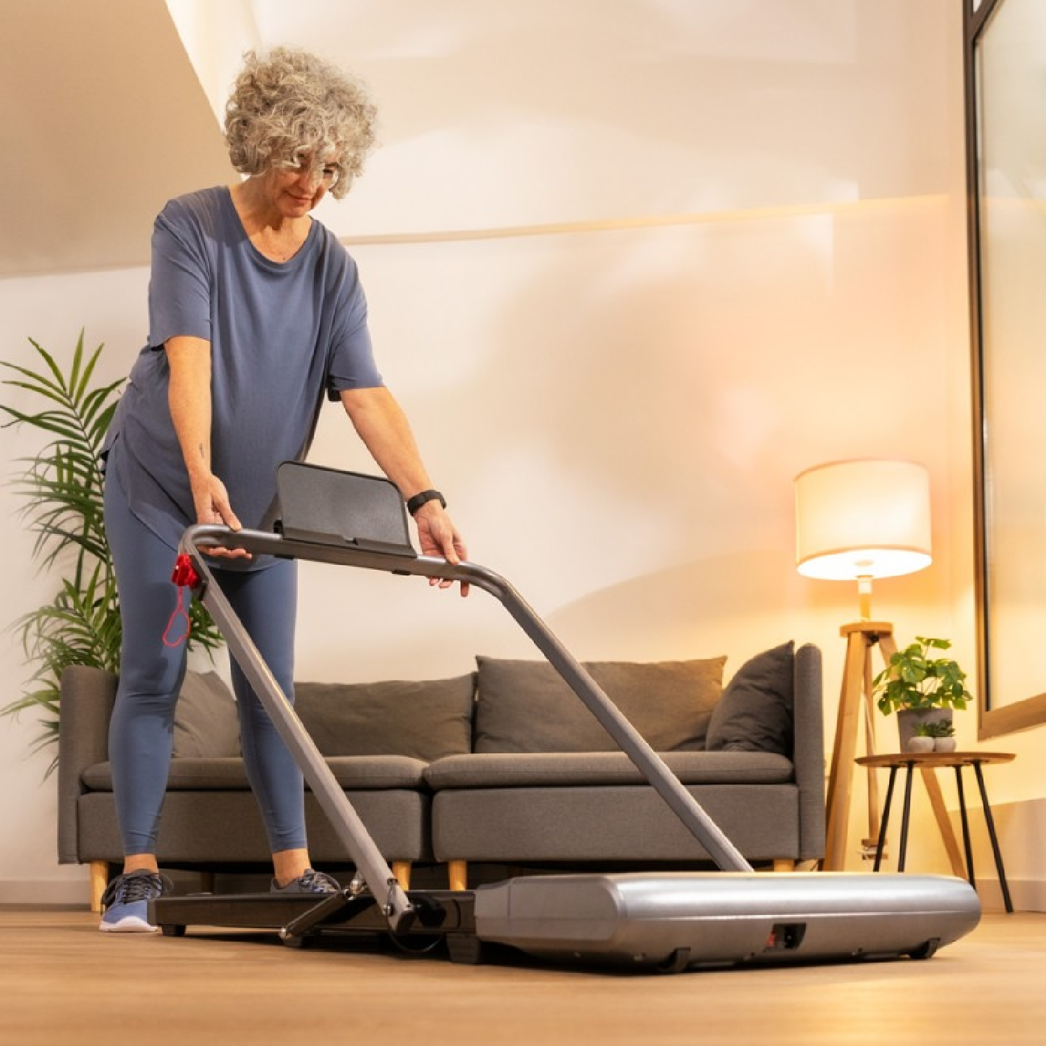 Starsza kobieta ustawiająca bieżnię w salonie. Neutralne odcienie.