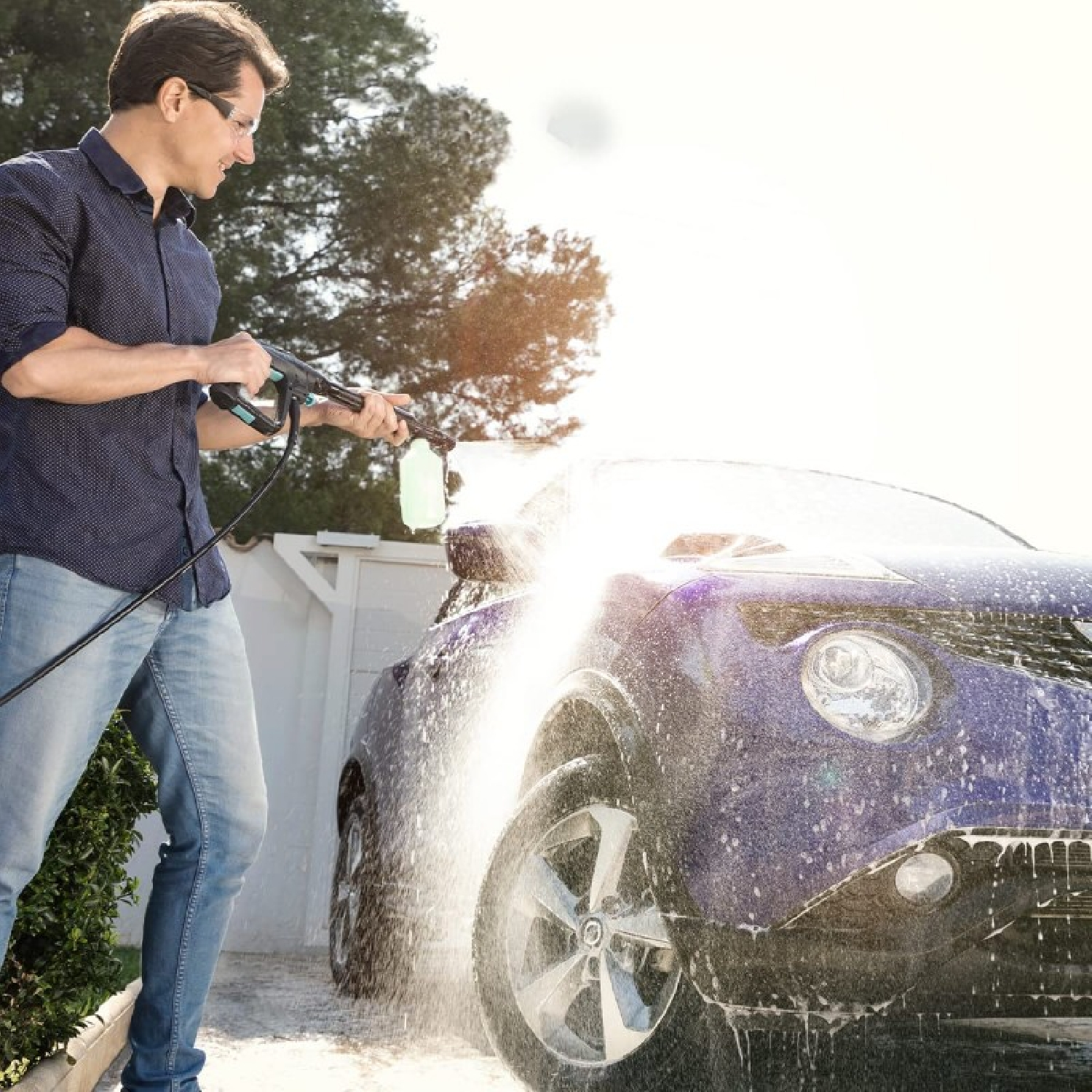 Uomo che lava un'auto blu con un'idropulitrice, creando uno spruzzo schiumoso.