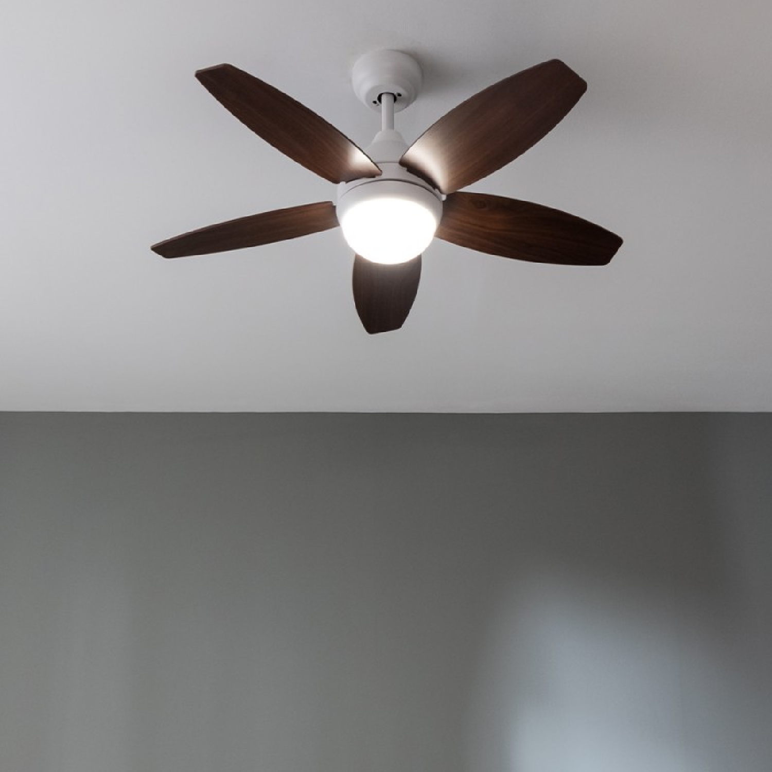 Ventilador de techo con cinco aspas de madera y luz central. Techo blanco y pared gris.
