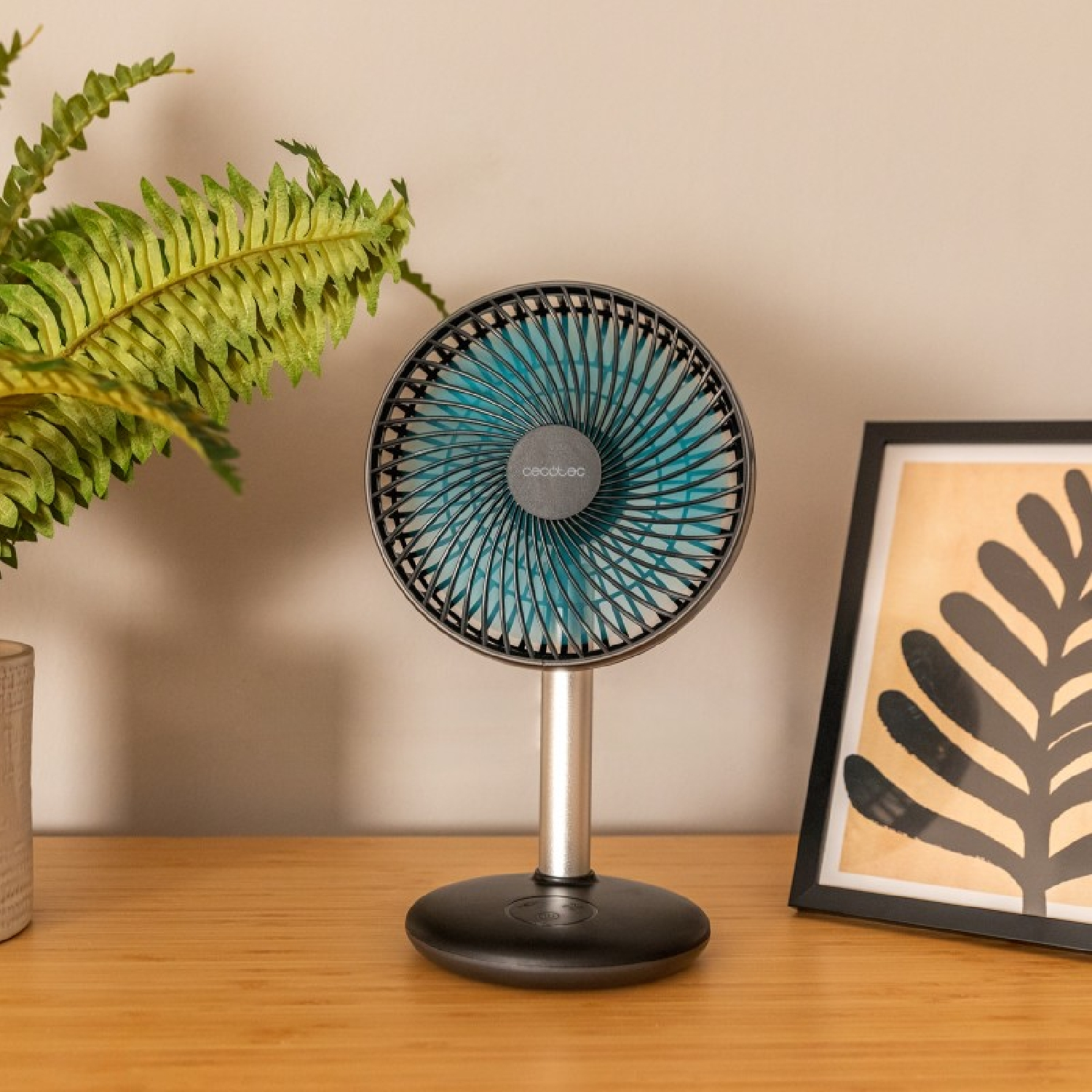 Un piccolo ventilatore su un tavolo di legno, con una felce e un'immagine incorniciata sullo sfondo.