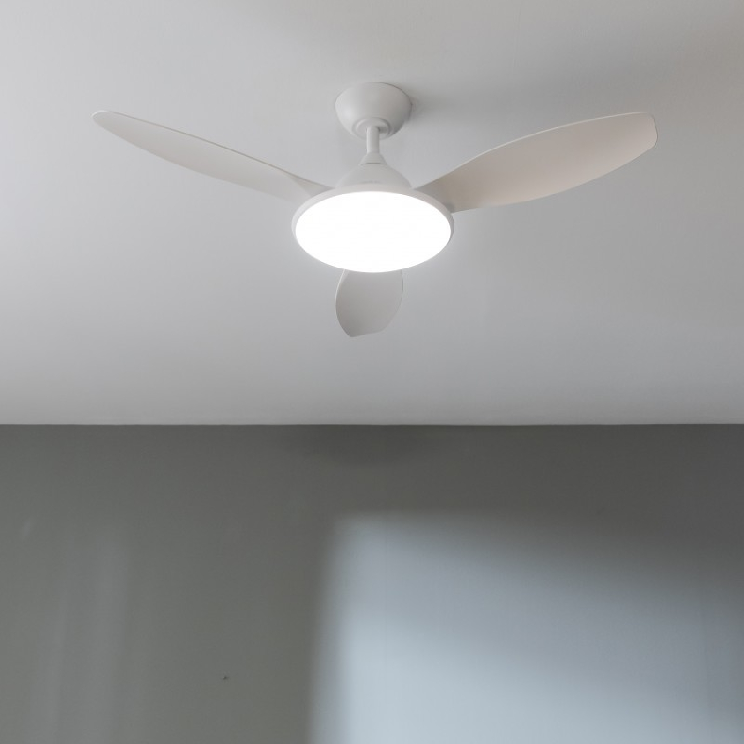 Un ventilatore a soffitto con tre pale bianche e una luce bianca. La ventola è contro un soffitto bianco.
