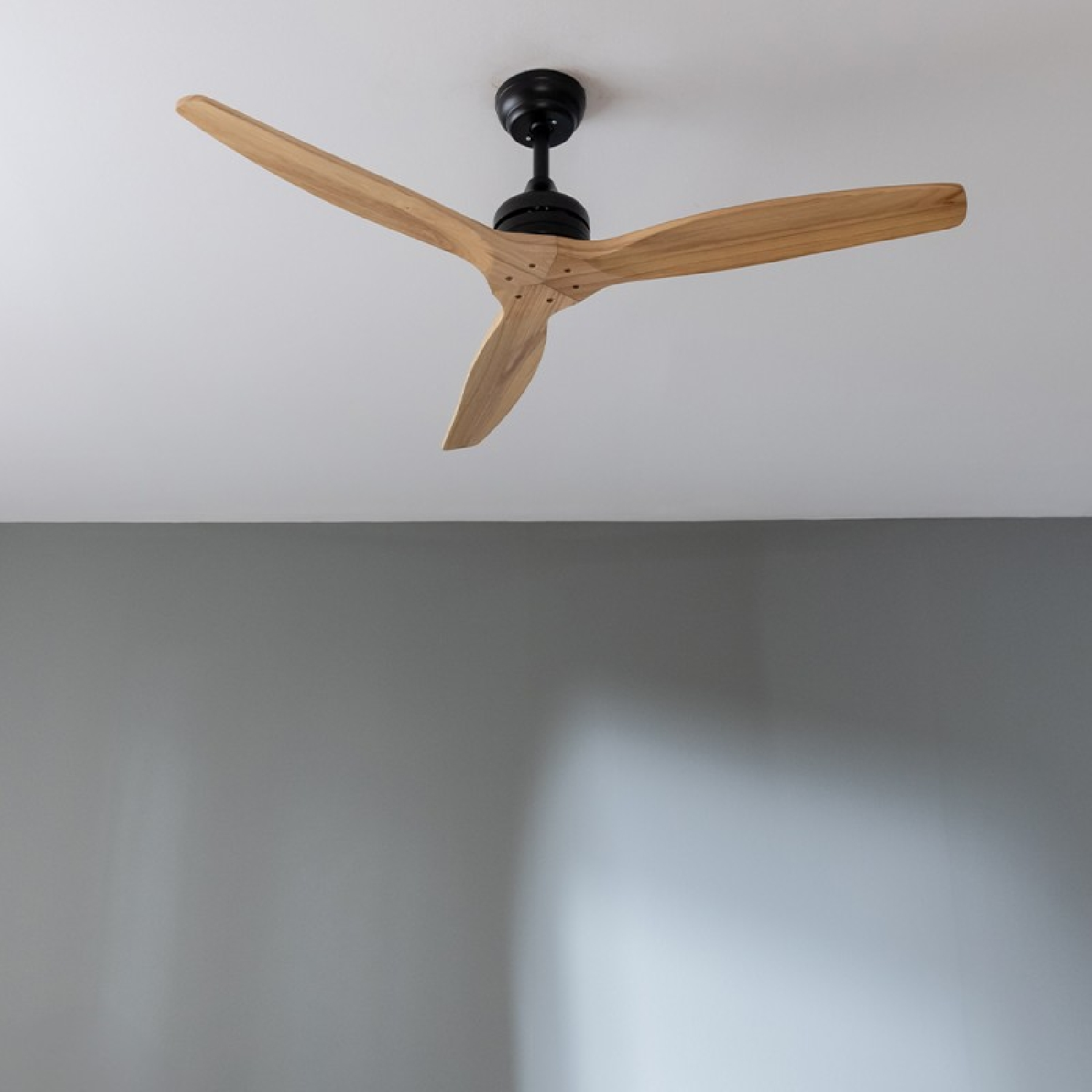 Deckenventilator mit Holzflügeln und schwarzem Motor. Er ist vor einer grauen Wand.