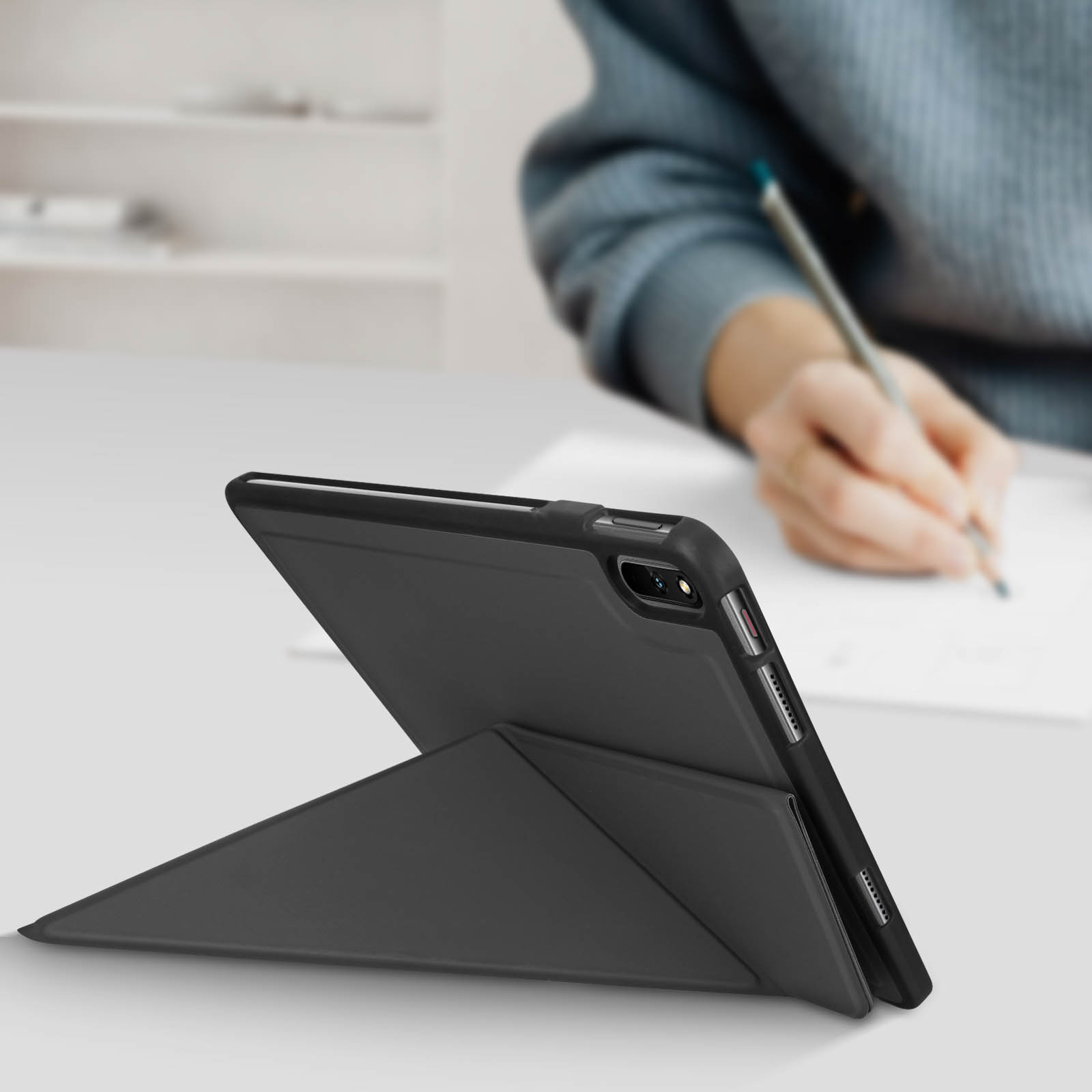 Tablet nero con supporto, angolato su una superficie bianca. Una persona scrive sullo sfondo.