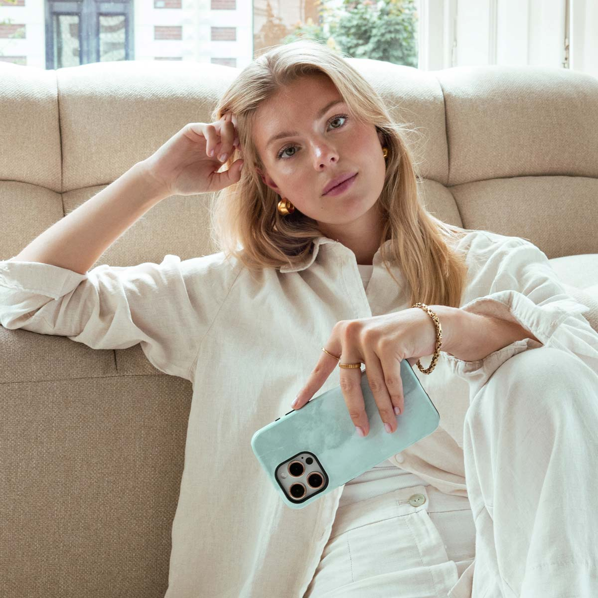 Blonde vrouw in witte outfit, houdt mintgroene telefoonhoes vast, zit op beige bank.