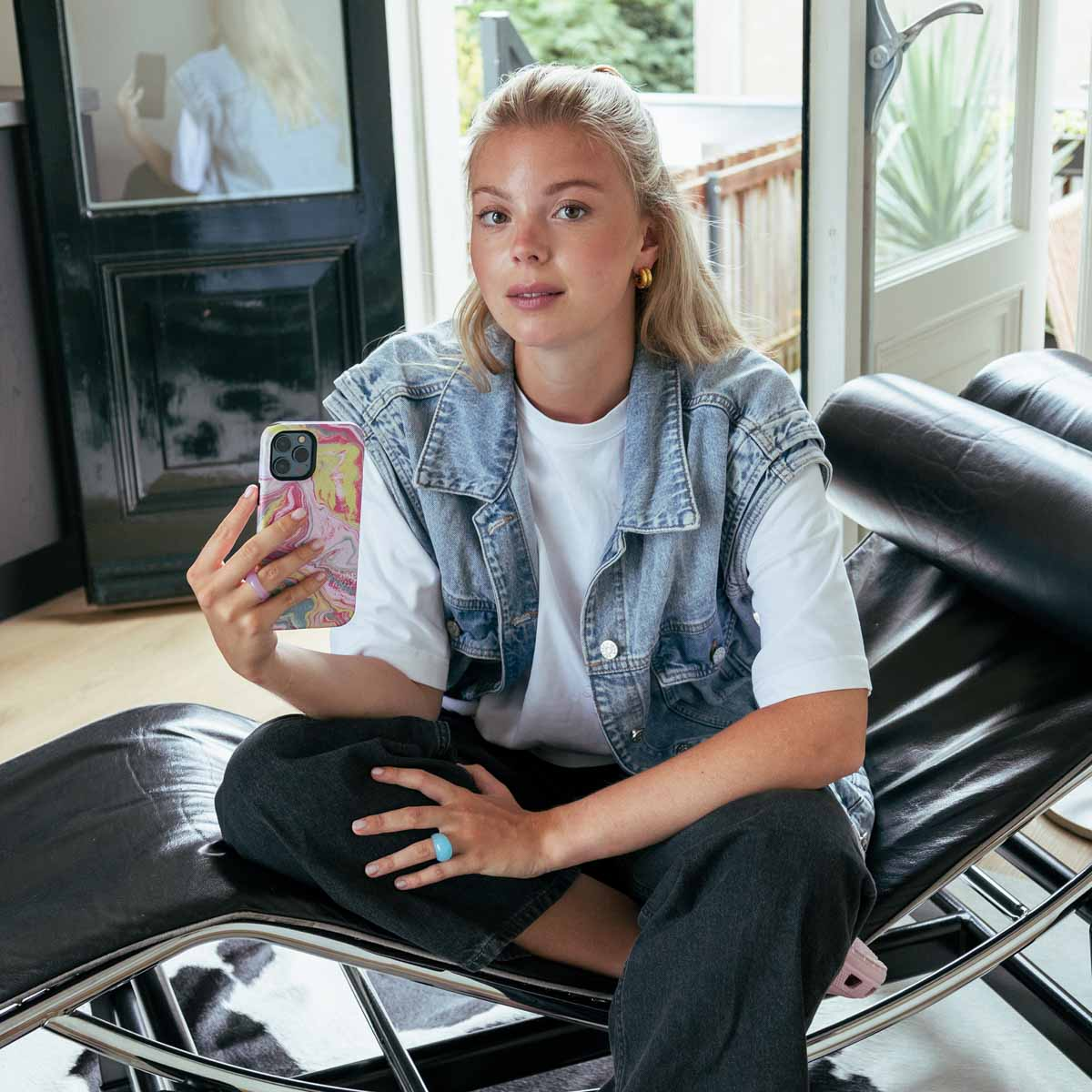 Une jeune femme blonde est assise sur une chaise noire, tenant un téléphone. Elle porte un gilet en jean et une chemise blanche.