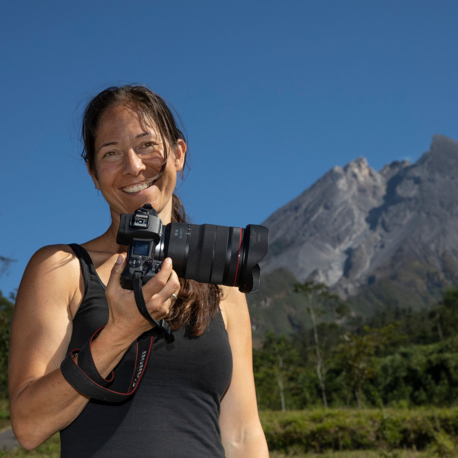 Frau lächelt, hält Kamera, Berg und blauer Himmel im Hintergrund.