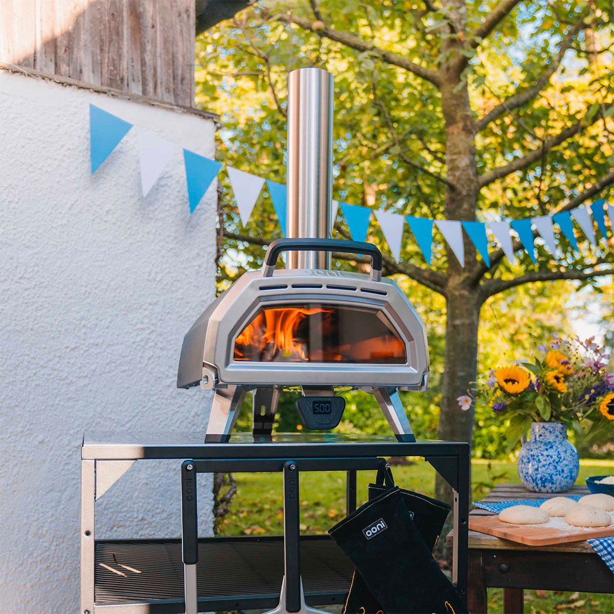 Forno per pizza all'aperto con fiamme, su un tavolo. Bandiere e alberi sullo sfondo.