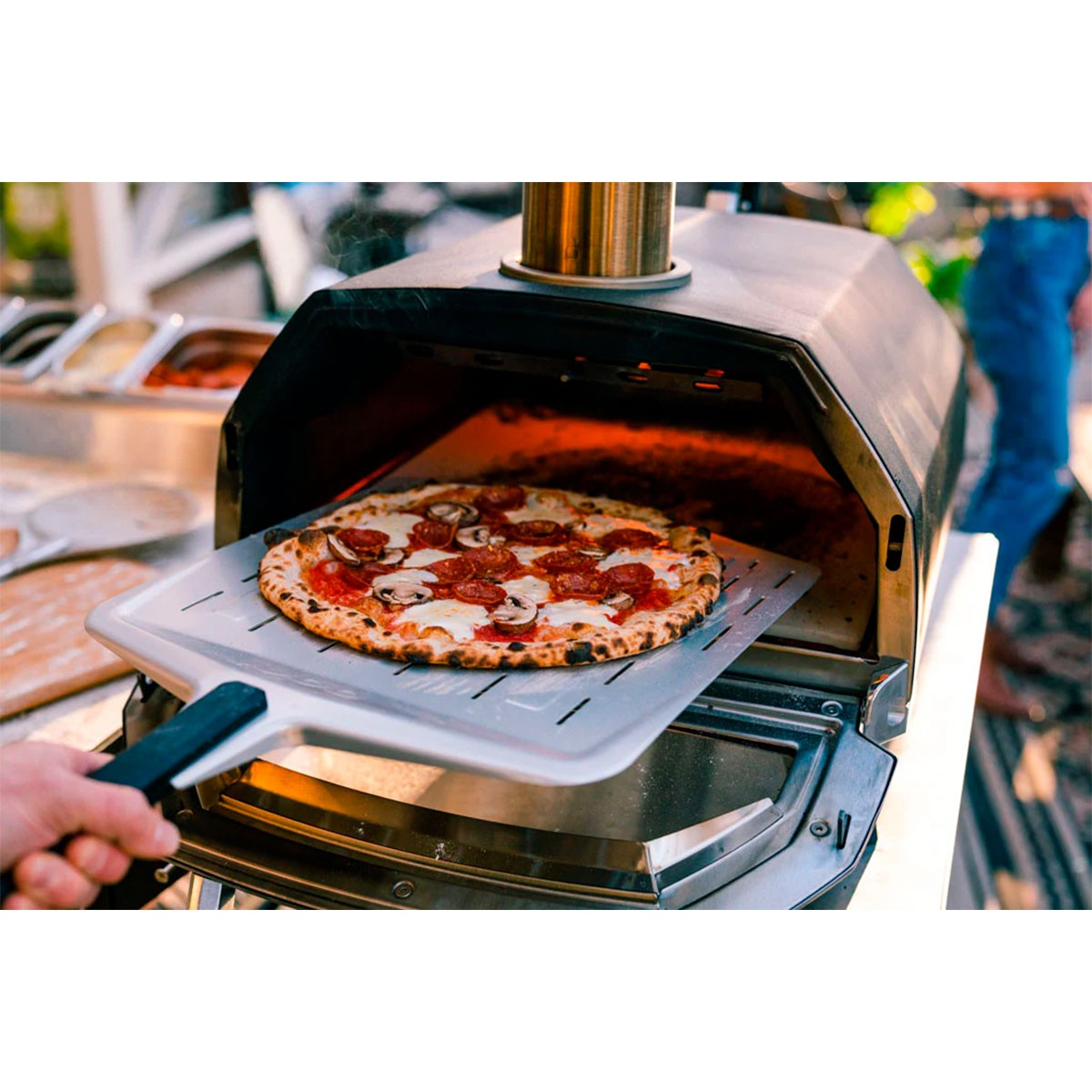 Pizza su una pala di metallo rimossa da un forno per pizza. Persona visibile sullo sfondo.