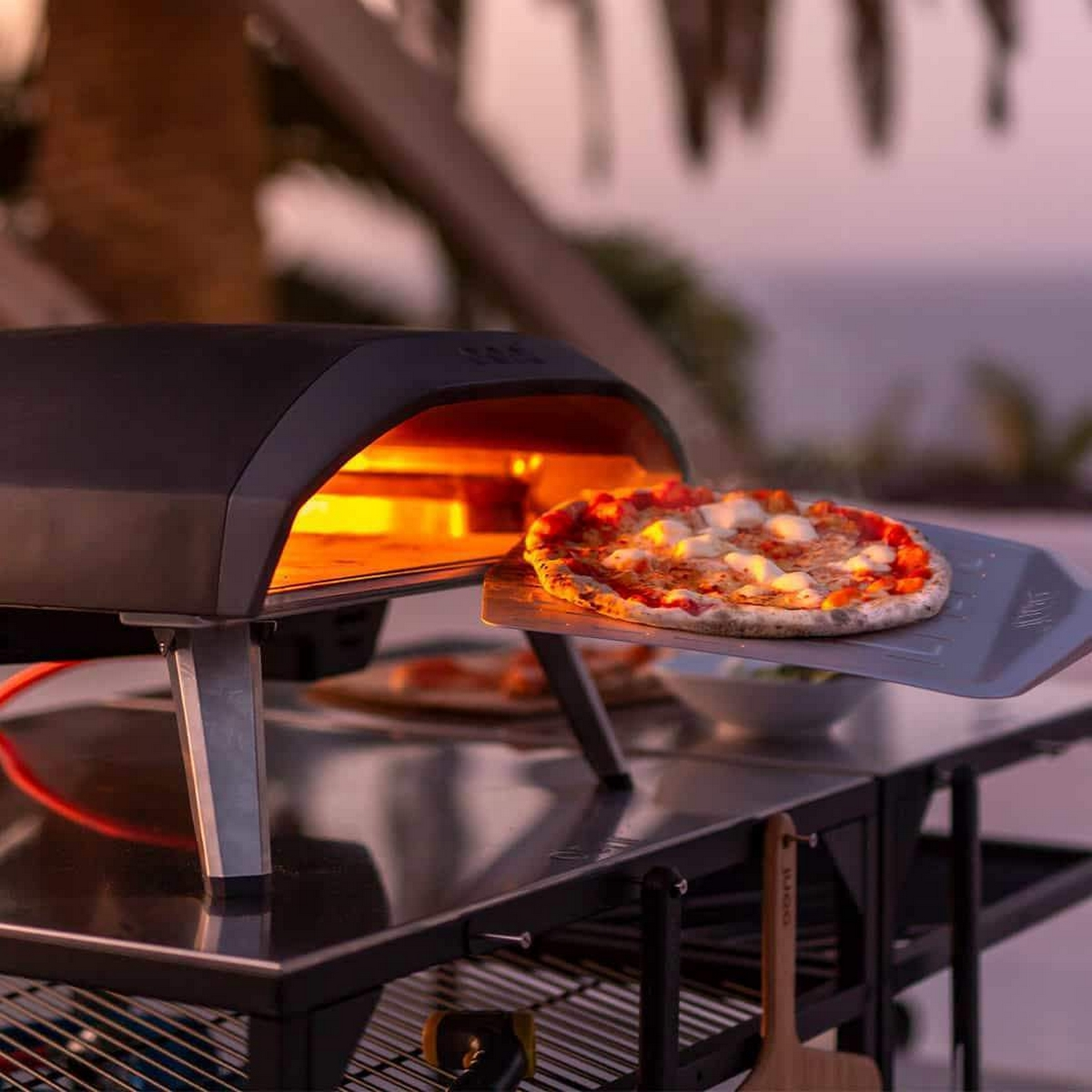 Una pizza viene rimossa da un forno per pizza incandescente con una pala; ambiente esterno.