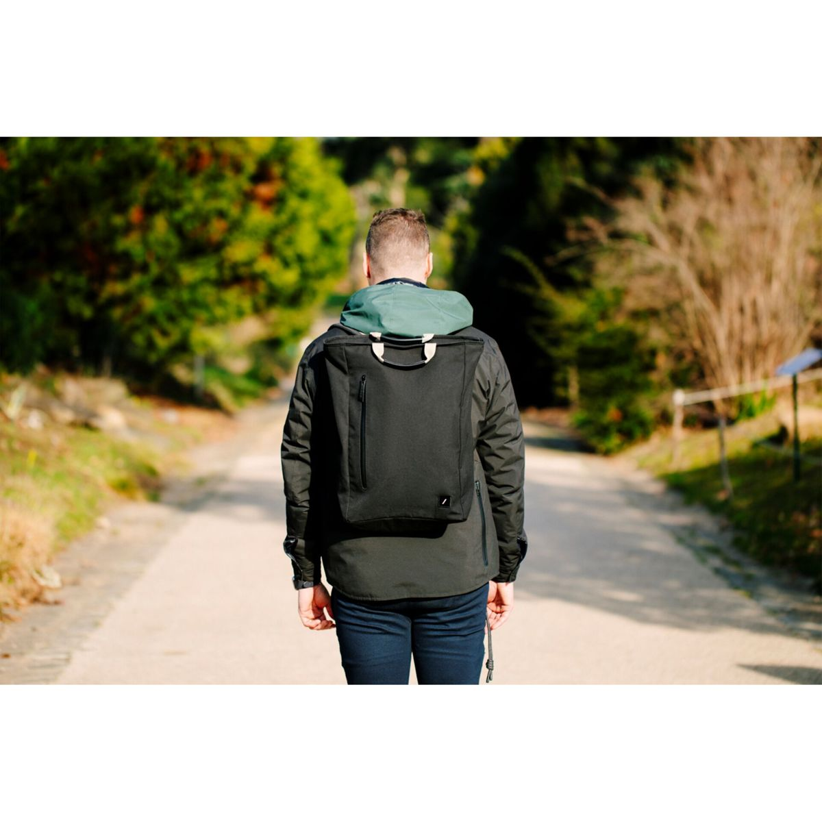 Mann mit schwarzem Rucksack geht auf einem Weg. Bäume säumen die Seiten. Die Person hat den Rücken zum Betrachter.