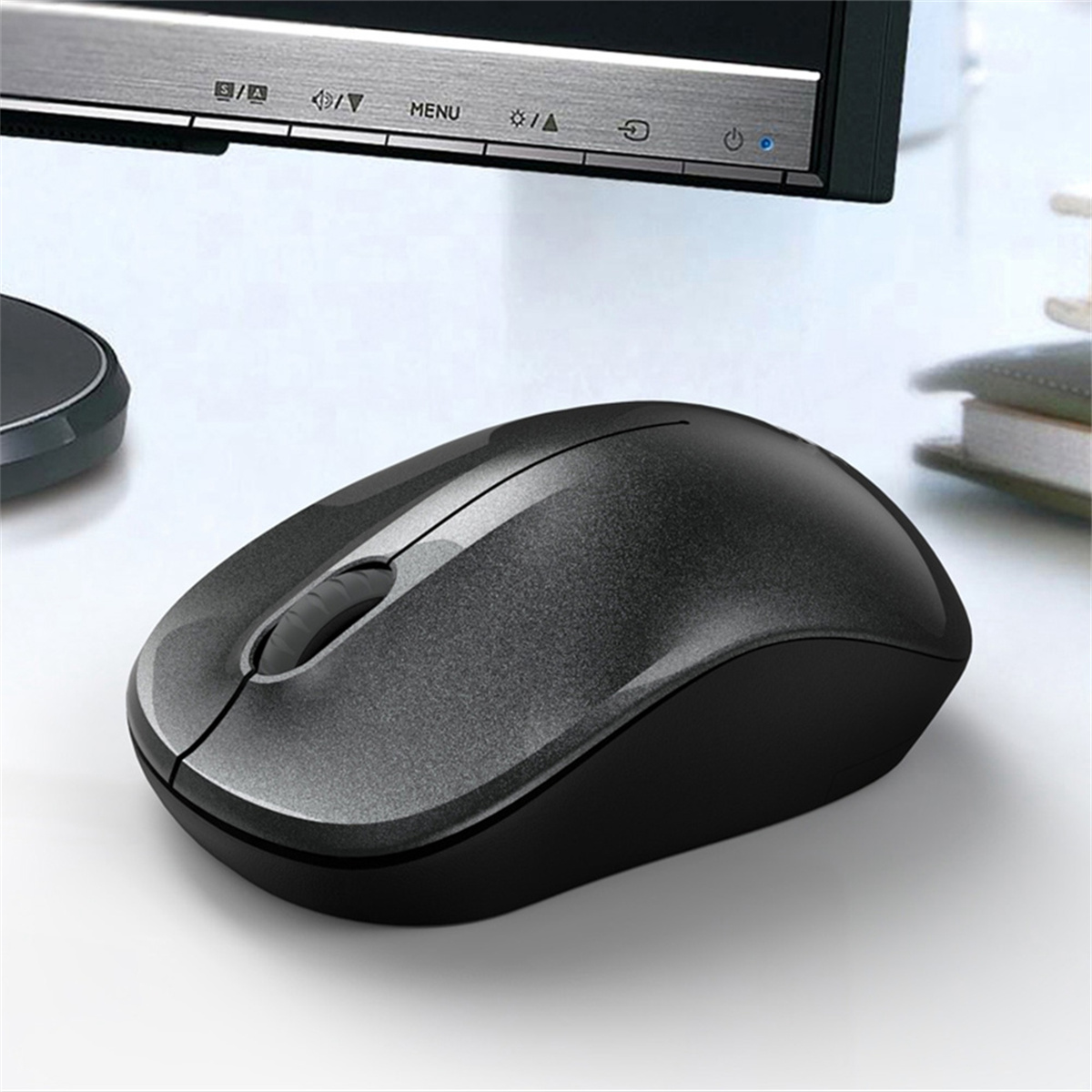 Eine grau-schwarze Computermaus liegt auf einem weißen Schreibtisch, im Hintergrund ein Monitor.
