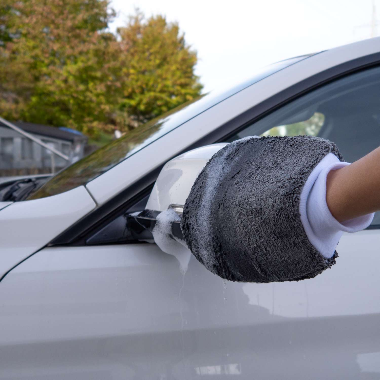 Eine Hand mit einem grauen Fäustling wäscht den Seitenspiegel eines weißen Autos. Der Spiegel ist mit Blasen bedeckt.
