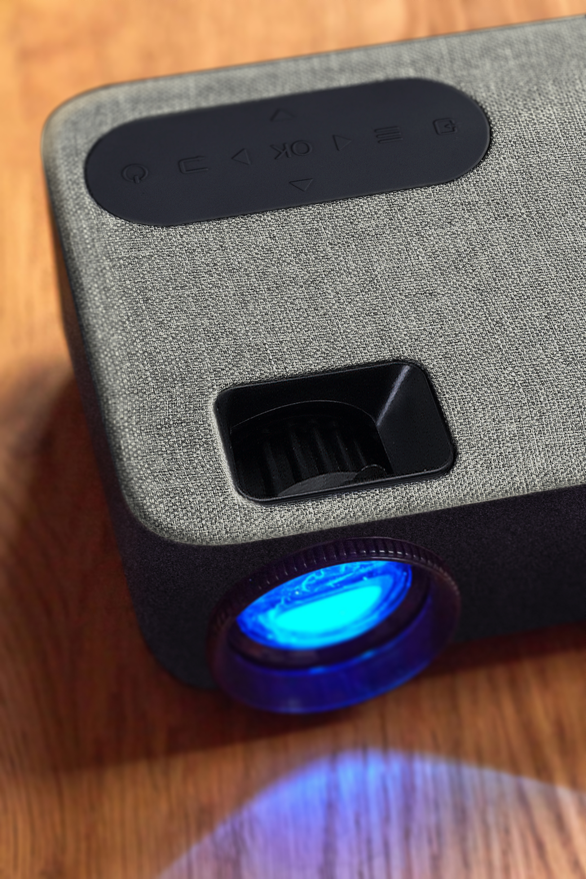 Szary projektor z obiektywem i panelem sterowania na drewnianej powierzchni.