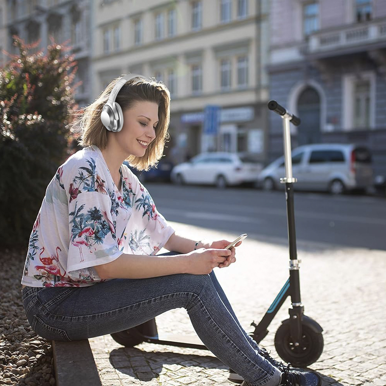 Donna con cuffie e telefono, seduta su un marciapiede, con uno scooter accanto.