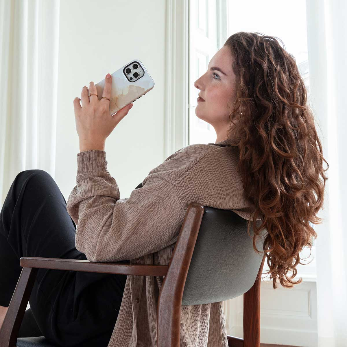 Vrouw houdt telefoon met hoes vast, zittend op een stoel. Krullend haar, neutrale tinten.